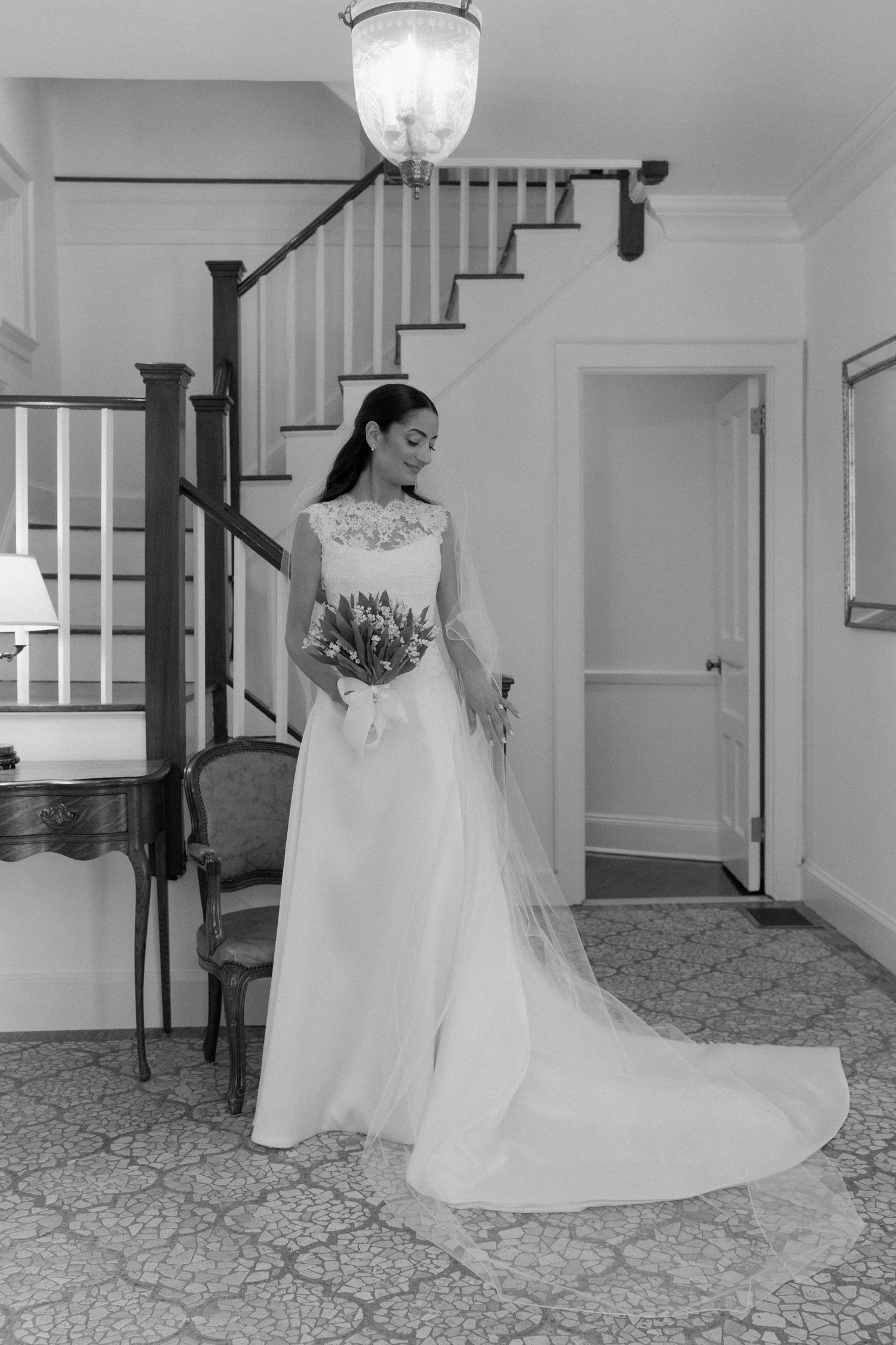 Elegant bride in a white lace gown holding a bouquet stands near a staircase in a classic interior.