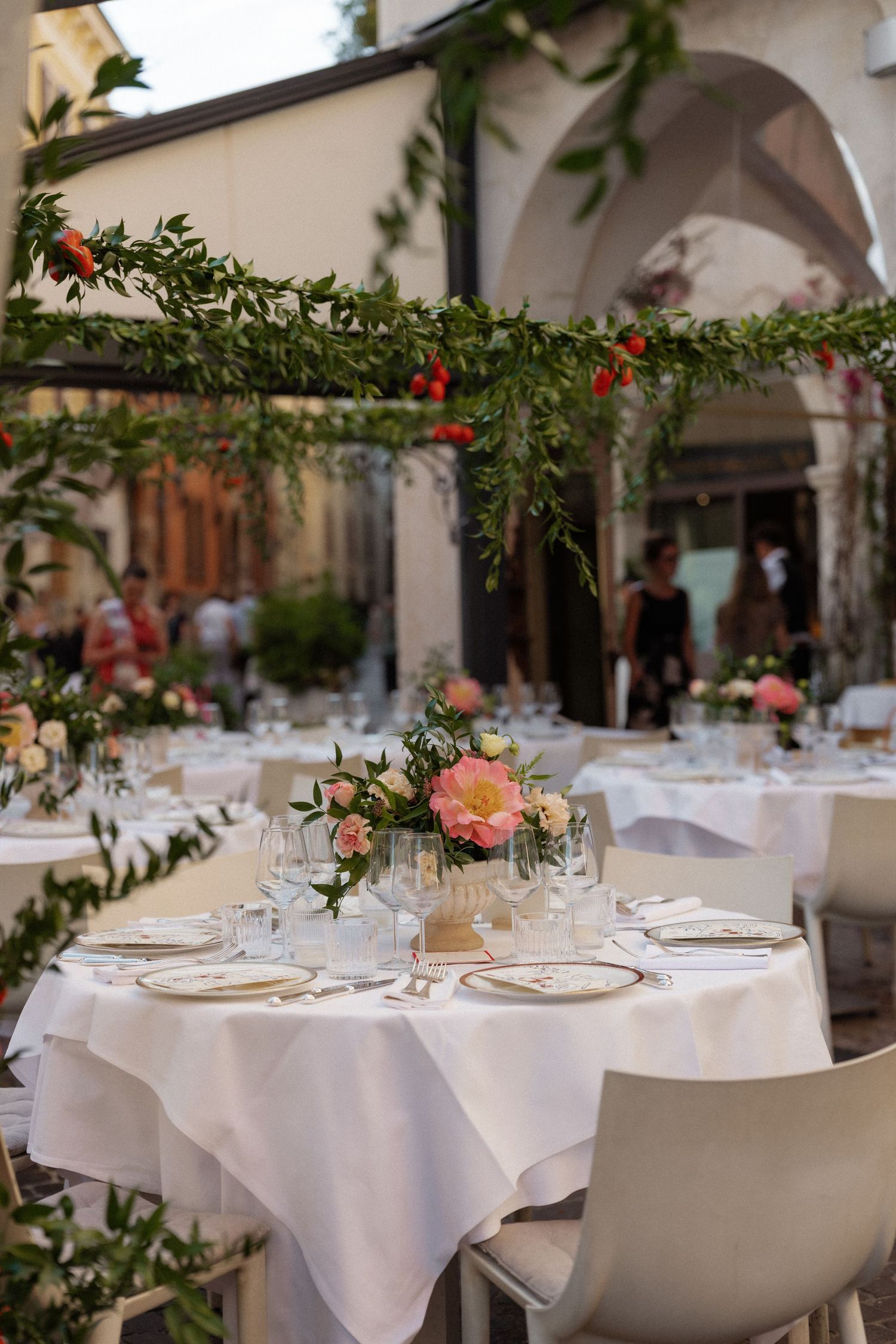 Elegant outdoor wedding reception tables with white linens, pink floral centerpieces, and greenery garland overhead decor.