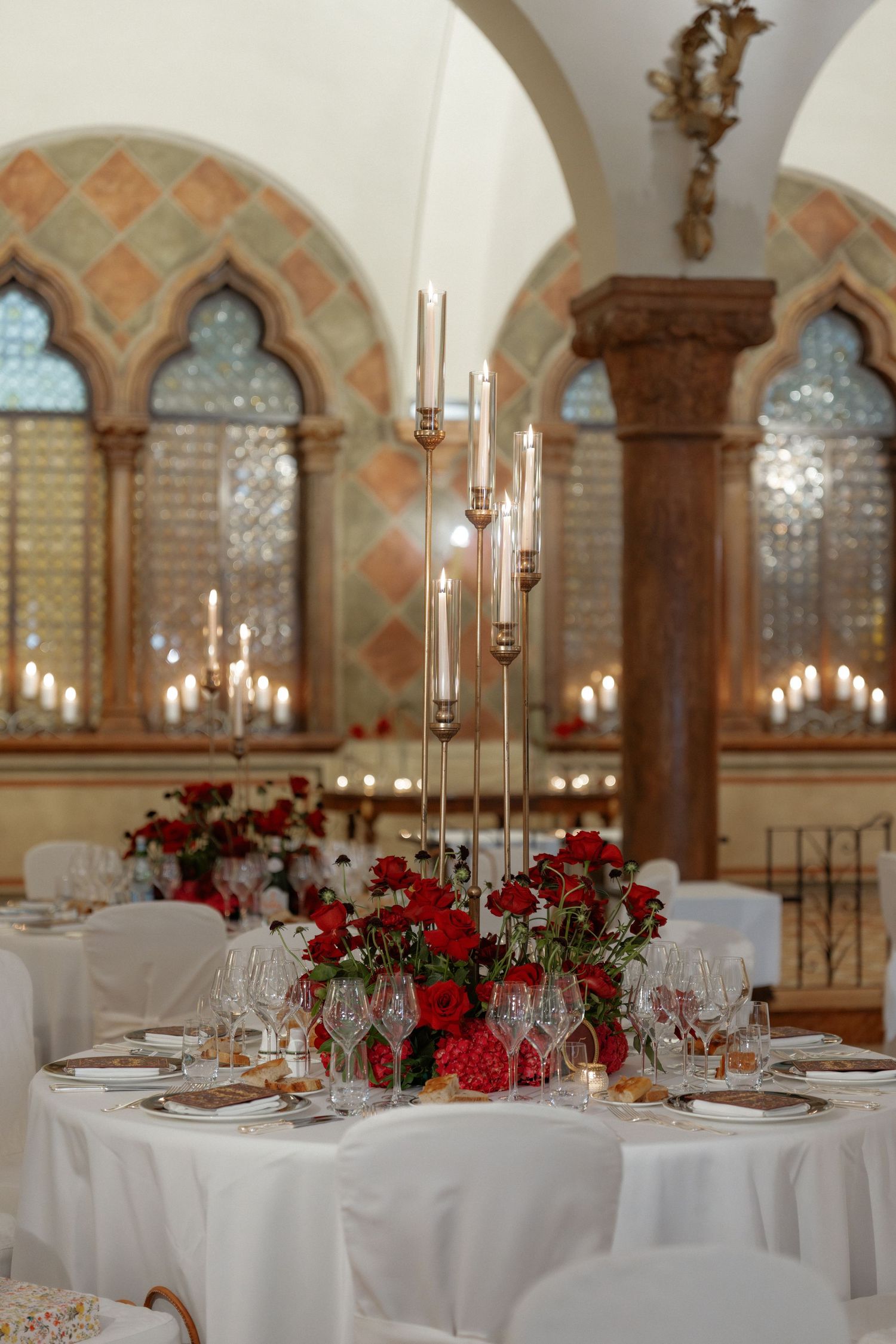 Elegant wedding reception hall with round tables, red floral centerpieces, tall candelabras, and arched windows.