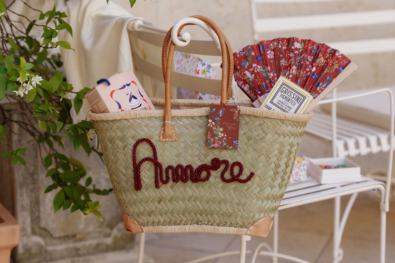 Woven straw tote bag with Amore embroidery, leather handles, filled with colorful accessories on a white chair.