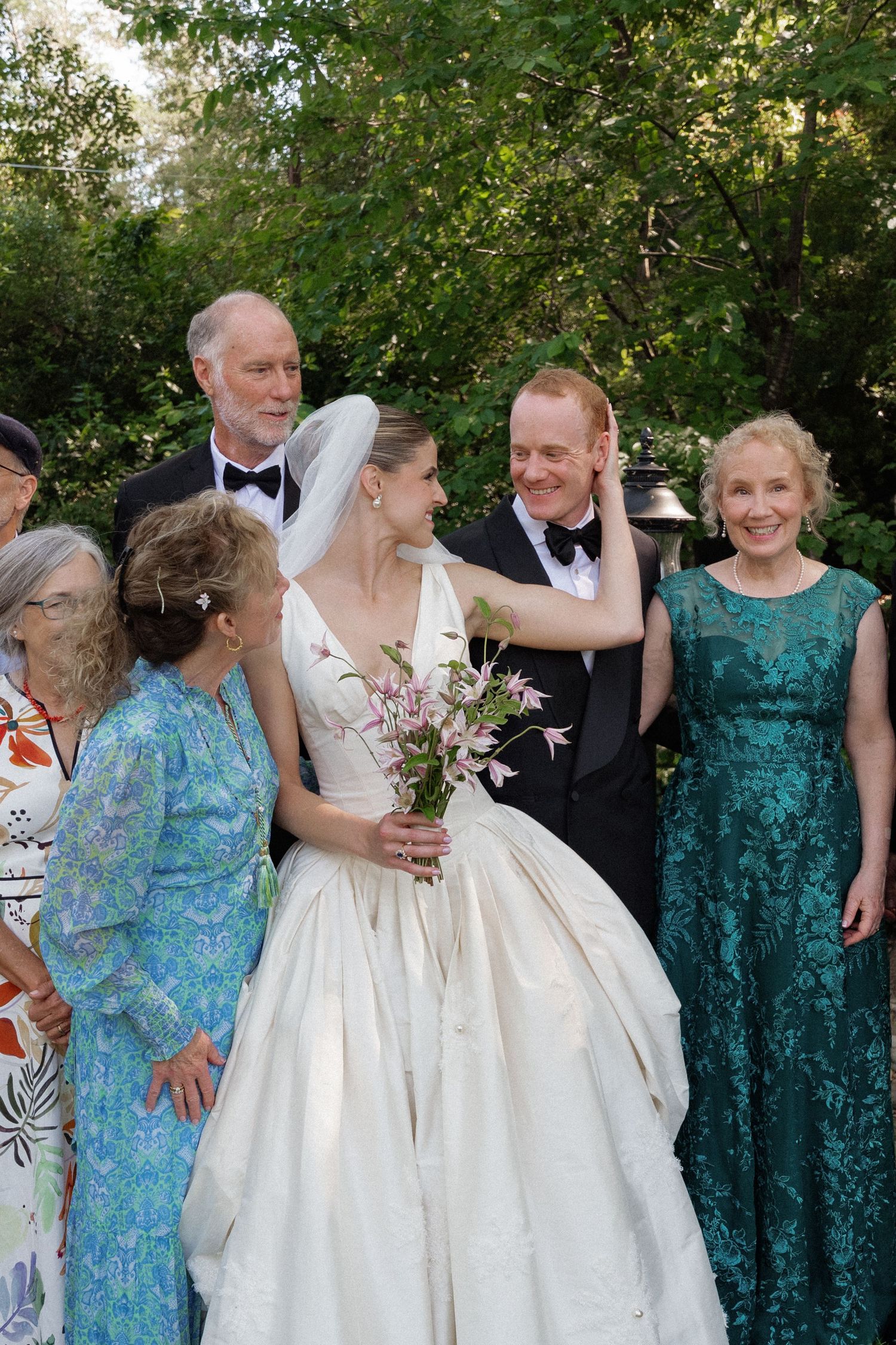 Bride in white gown holds pink flowers beside groom in tuxedo, surrounded by smiling family outdoors.