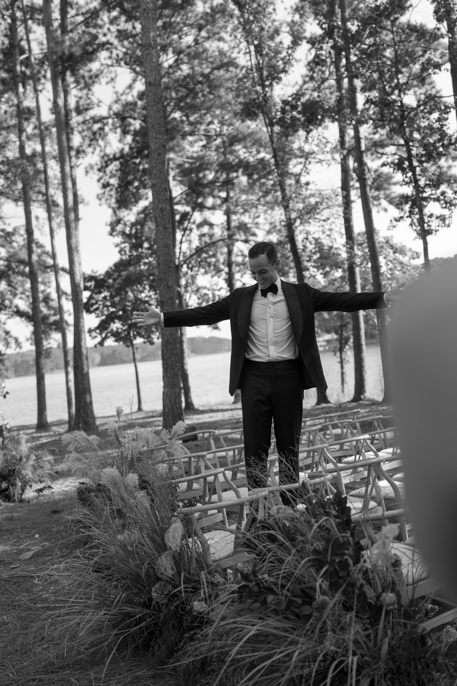 Wedding officiant with arms raised during outdoor ceremony among pine trees and wooden benches.