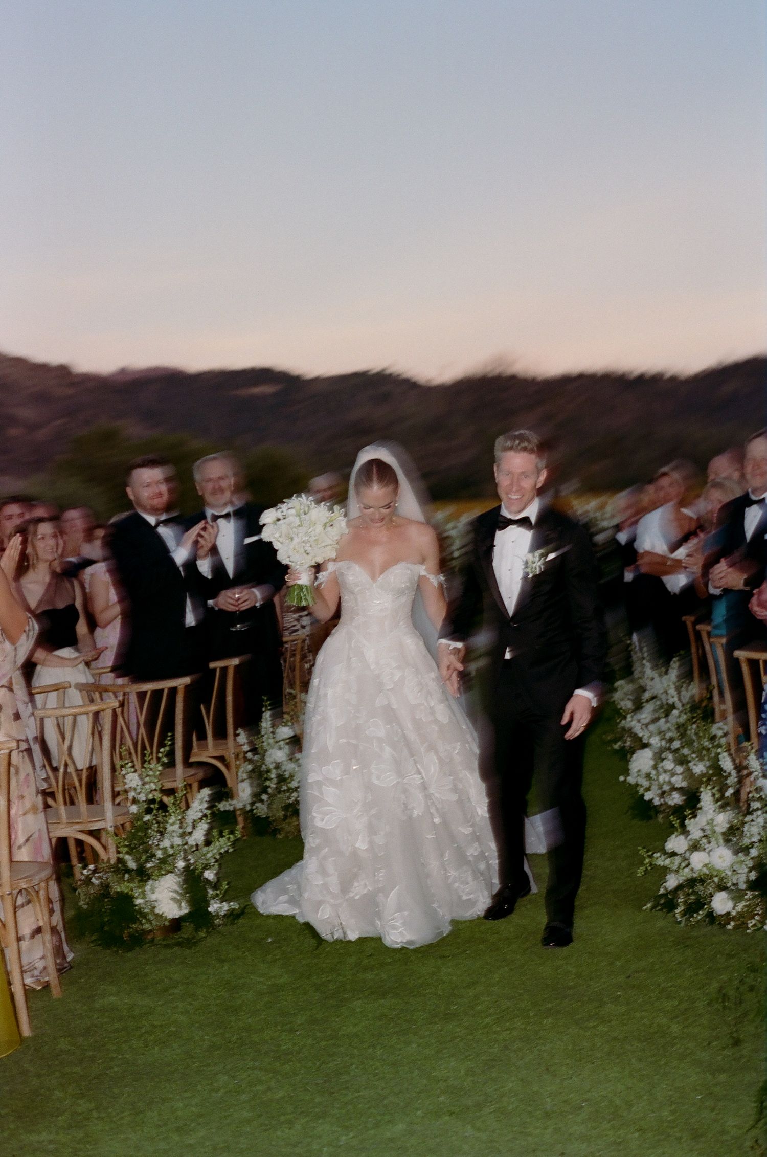 Outdoor evening wedding ceremony with guests seated on wooden chairs among floral decorations.