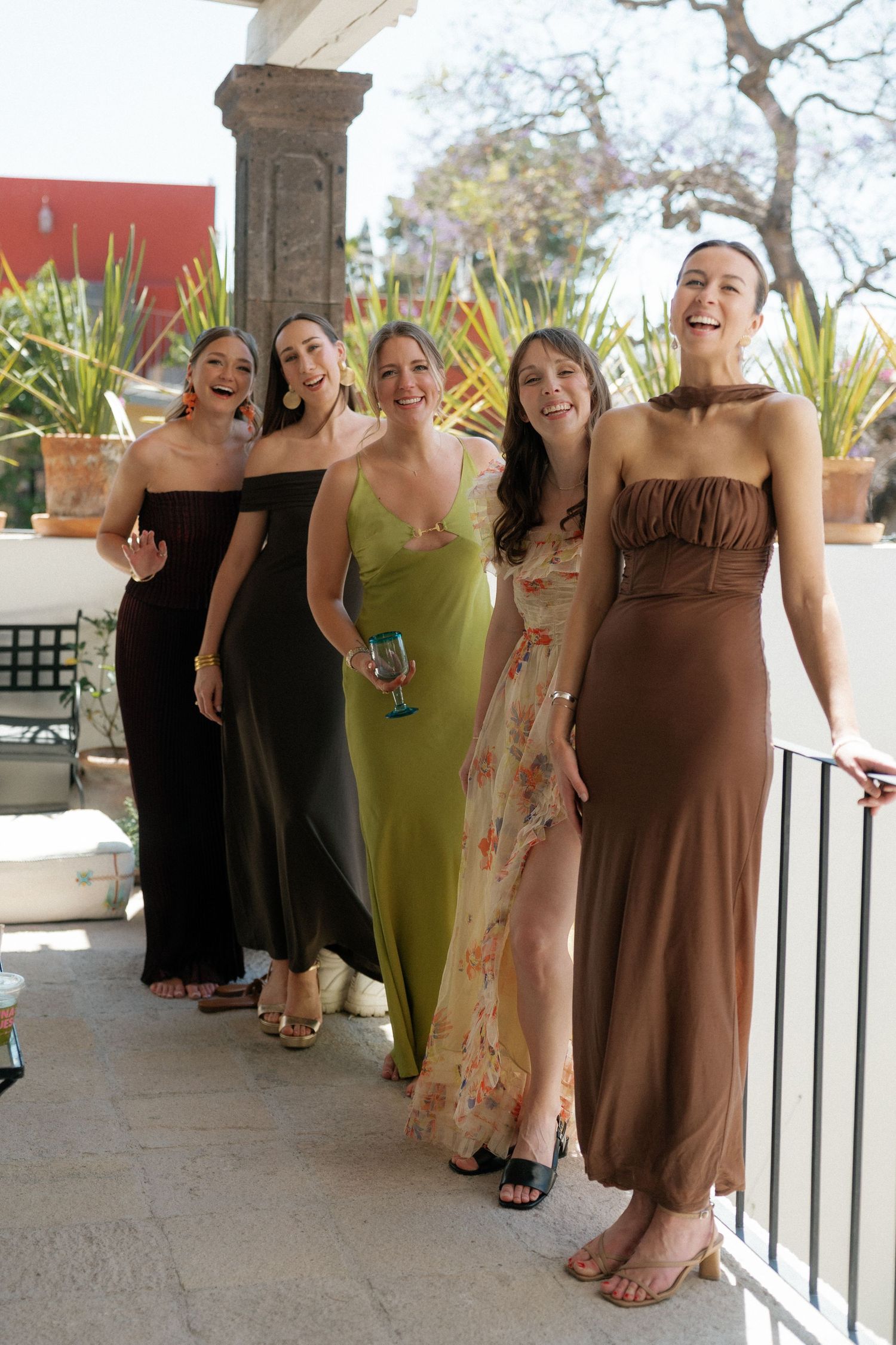 Bridesmaids in brown and green dresses posing together on an outdoor terrace.