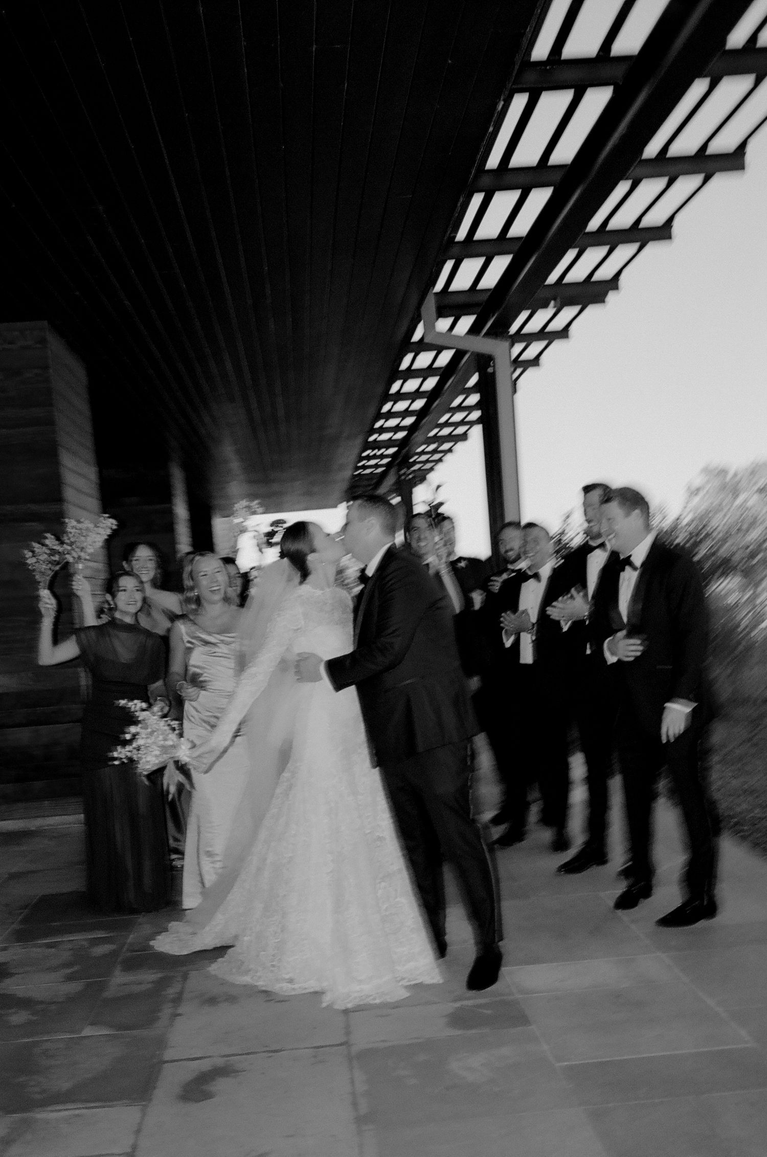 Black and white photo of wedding reception with guests dancing under architectural beams.
