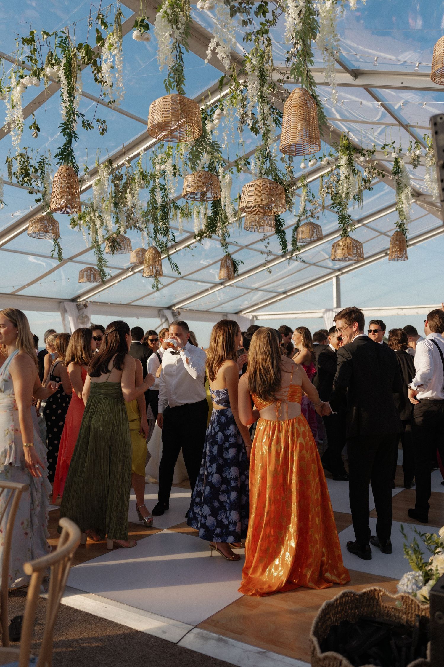 Elegant wedding reception under a clear tent adorned with hanging wicker lanterns and greenery, guests mingling in formal attire.