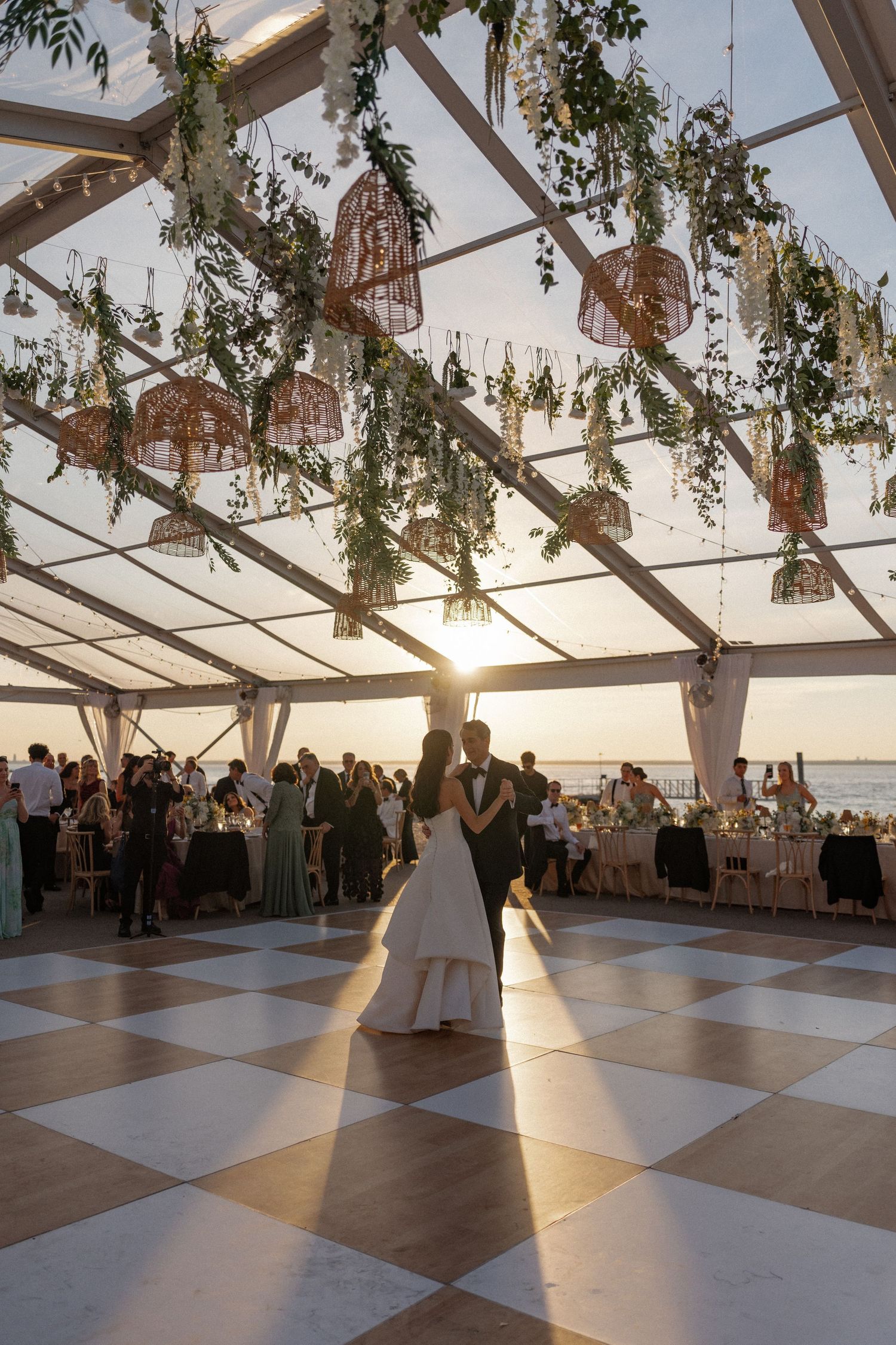 Bride and groom share first dance under a clear tent adorned with greenery and lanterns at a sunset waterfront wedding.
