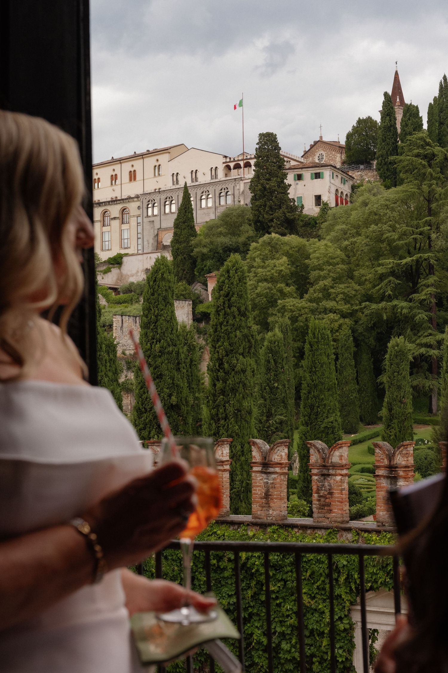 Woman holding aperol spritz on balcony overlooking Italian villa and cypress trees with Italian flag.