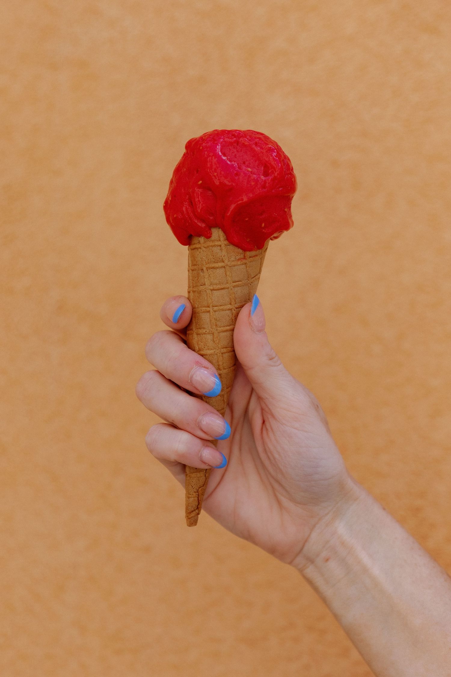 Hand with blue nail polish holding a red strawberry gelato scoop in a waffle cone against an orange wall.
