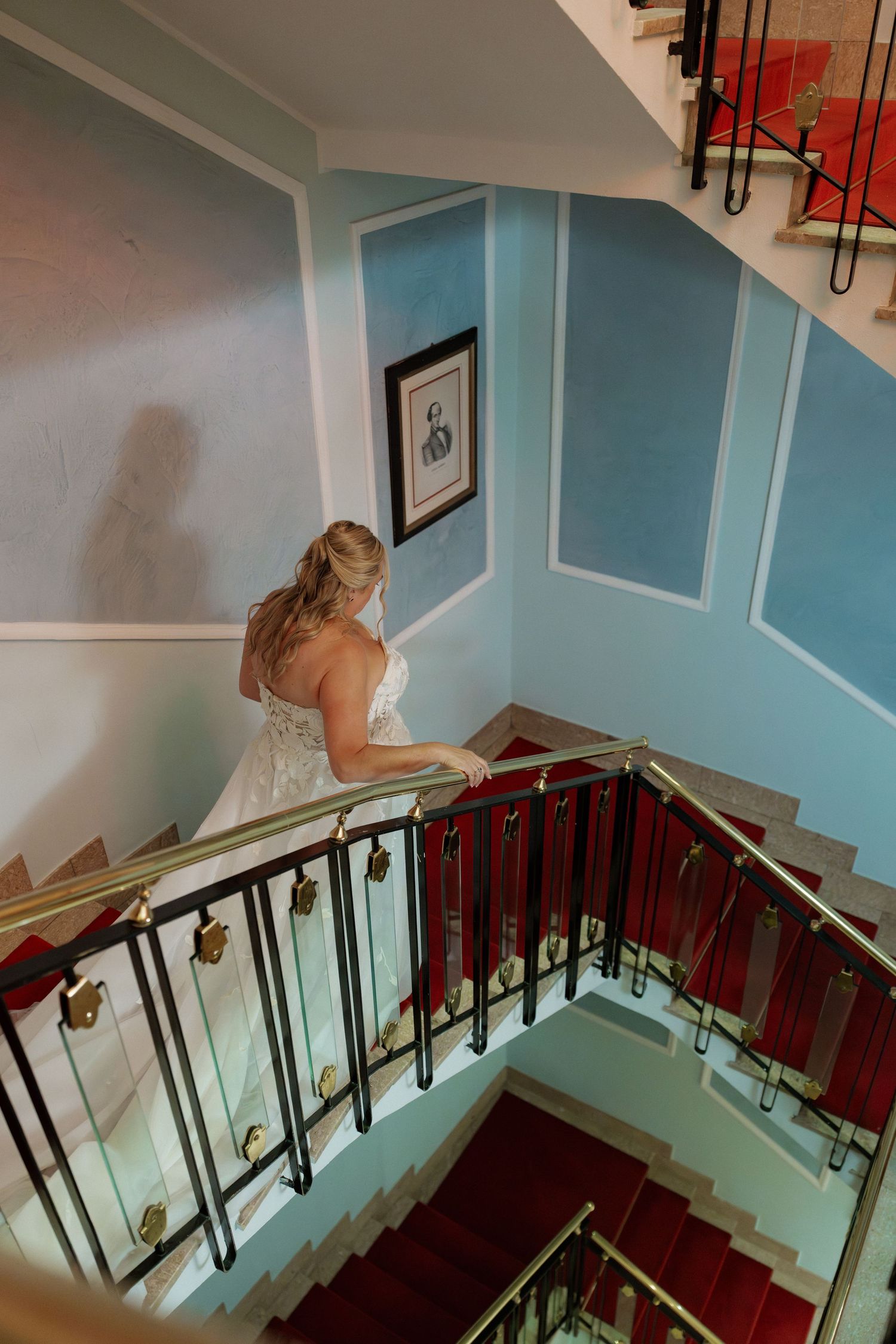 Bride in white lace wedding gown descending elegant staircase with red carpet and gold railings in blue-walled venue.