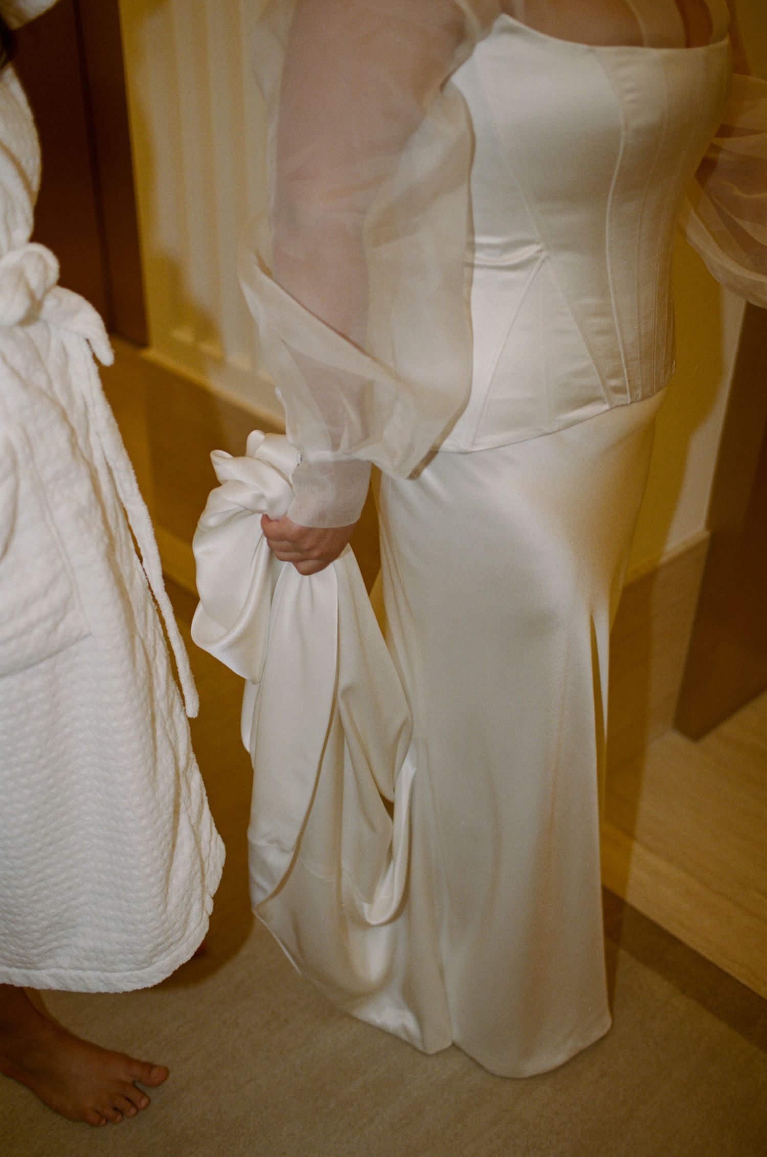 Close-up details of a flowing white wedding dress and bare feet on a carpeted floor.