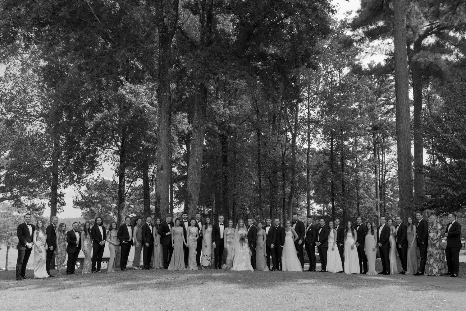 A black and white wide shot of a wedding party lined up among tall trees.