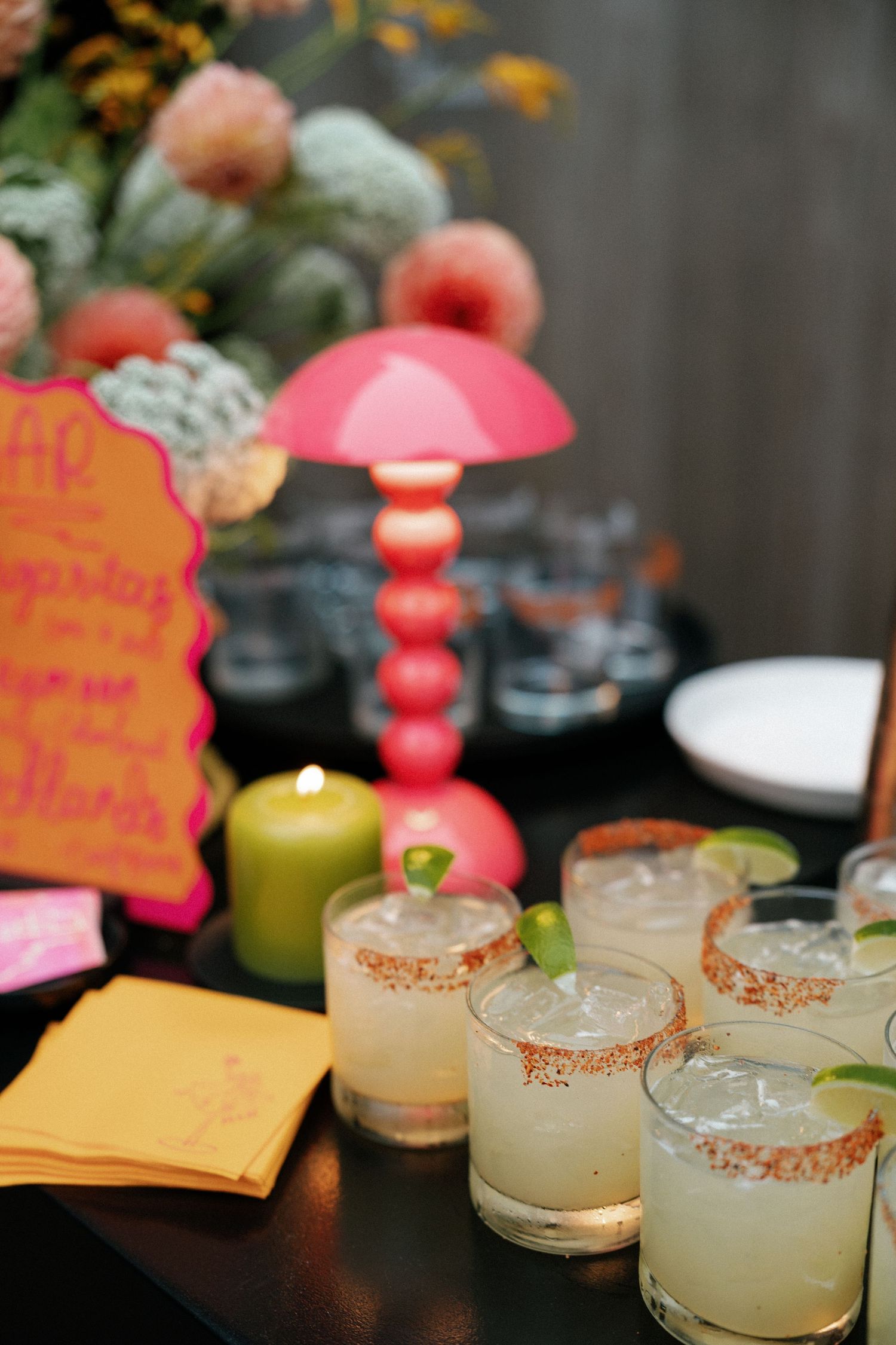Elegant cocktail glasses with salt rims and orange garnishes sit alongside decorative pink signs on a dark table.
