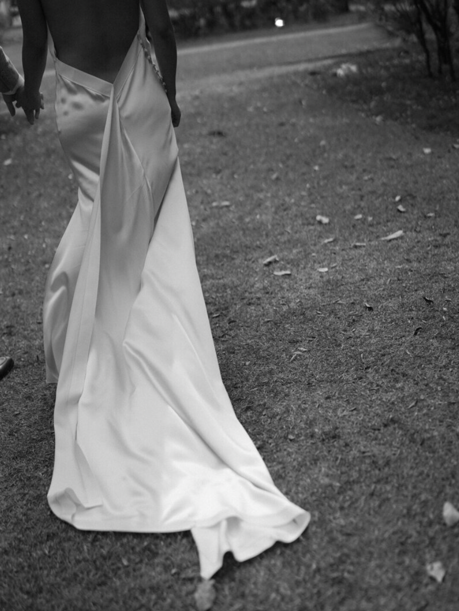 Close-up black and white photo of a flowing white wedding dress train on grass.