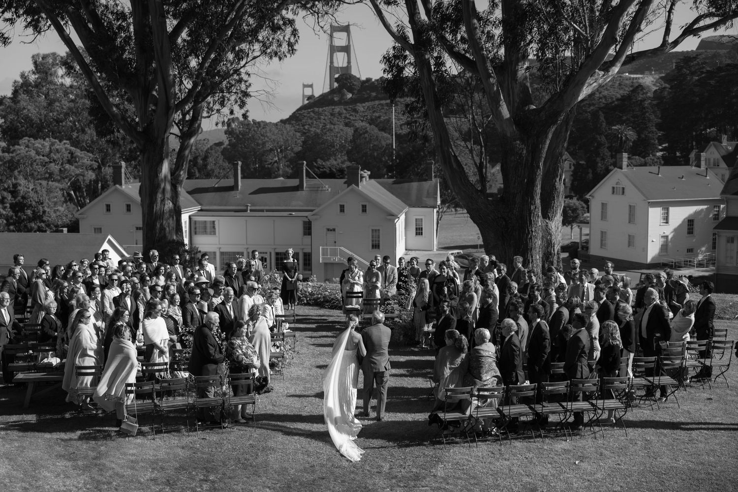 Black and white photo of outdoor wedding ceremony with Golden Gate Bridge visible through trees.