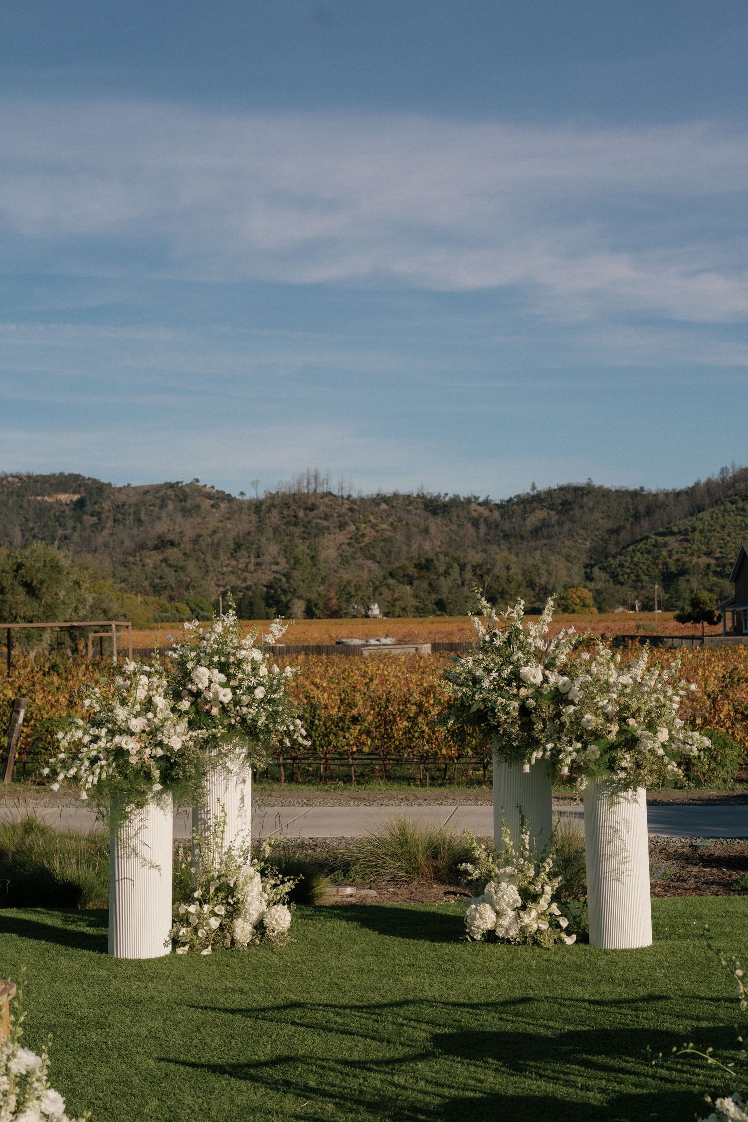 White floral arrangements in milk jug vases line a grassy outdoor wedding ceremony space.