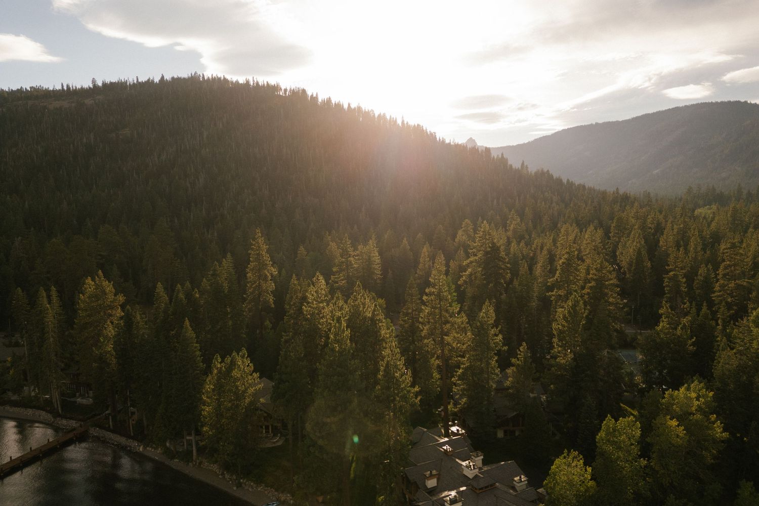 Golden sunlight breaks through clouds over a mountainous forest landscape at sunset.