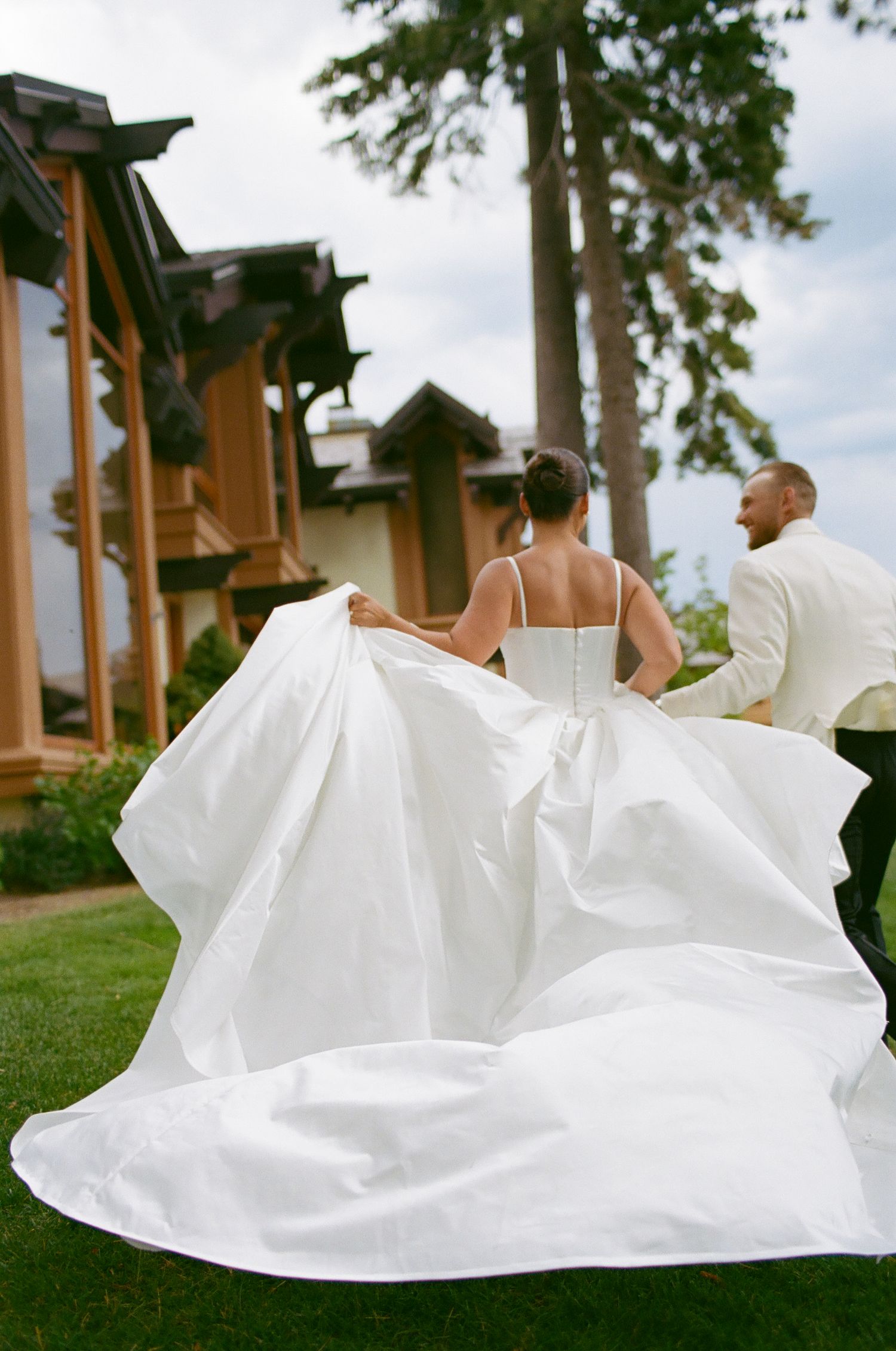 A flowing white wedding dress billows in the breeze as a couple walks on a lawn near wooden buildings.
