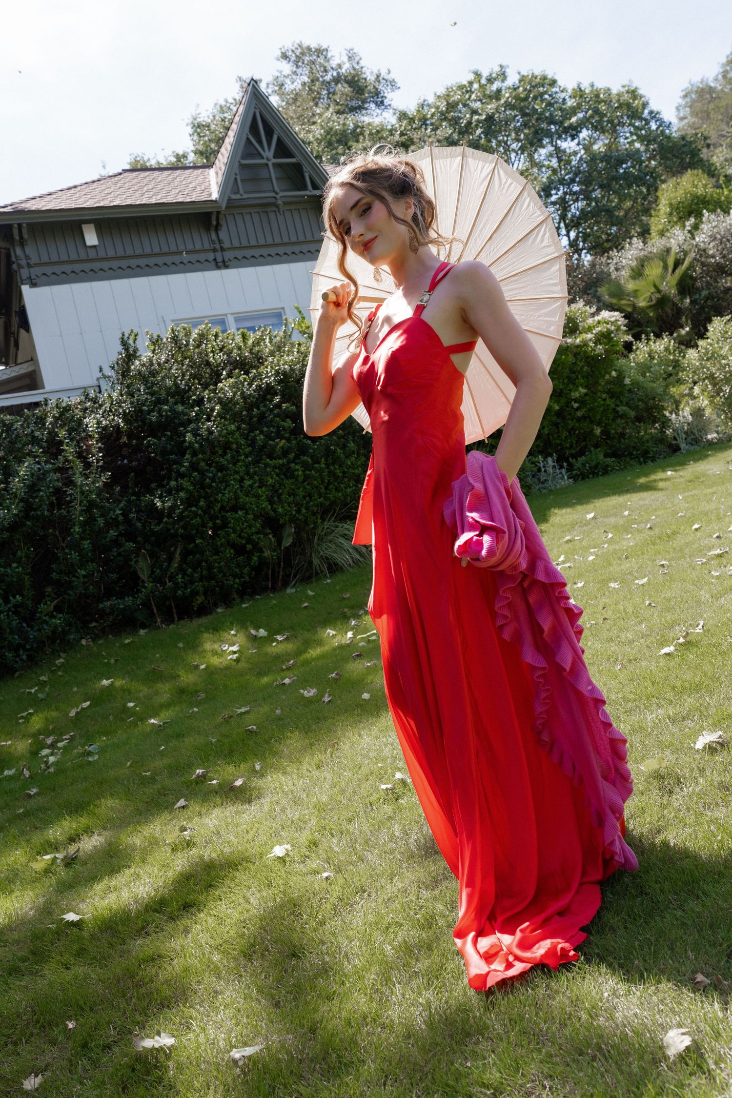 Woman in elegant red gown holding parasol poses on green lawn with garden backdrop.