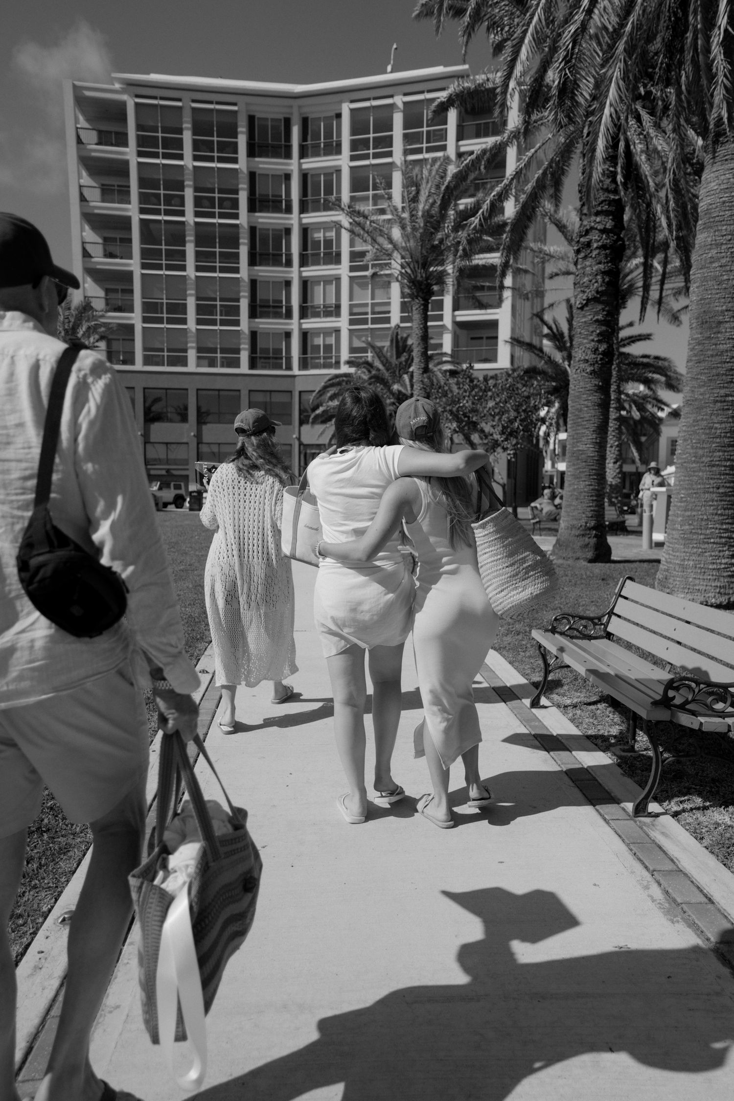 A black and white photograph showing people walking on a sidewalk in front of a large building with palm trees.