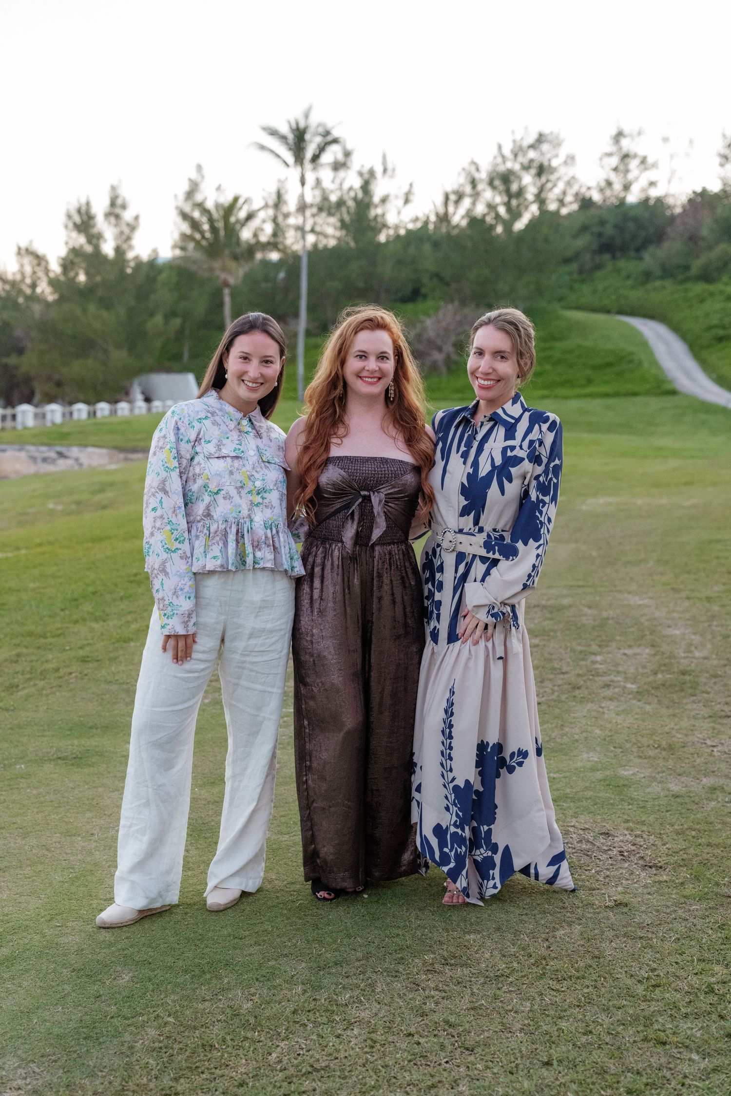 Three young women in colorful patterned dresses stand together on a grassy field with trees in the background.