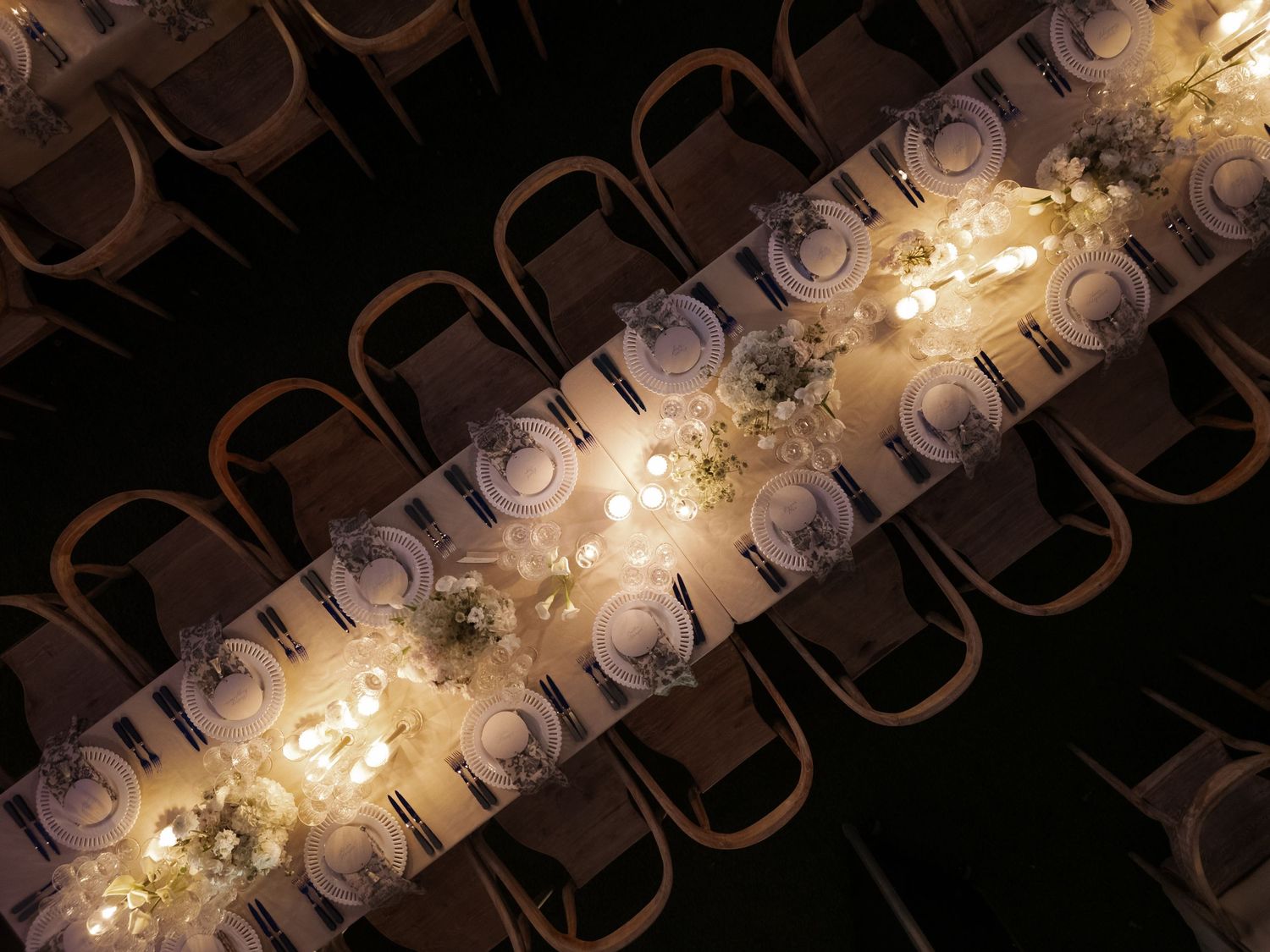 Elegant long dining table with glowing candles, white floral centerpieces, and place settings viewed from above in a dark venue.