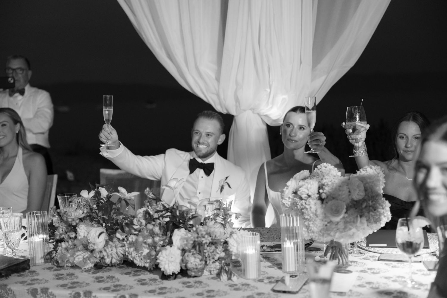 Wedding reception tables decorated with floral centerpieces and white draping in an elegant tent setting.