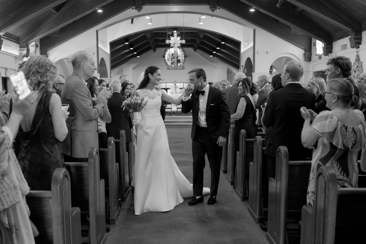 Black and white photo of newlyweds sharing a kiss at the church altar aisle, surrounded by guests.
