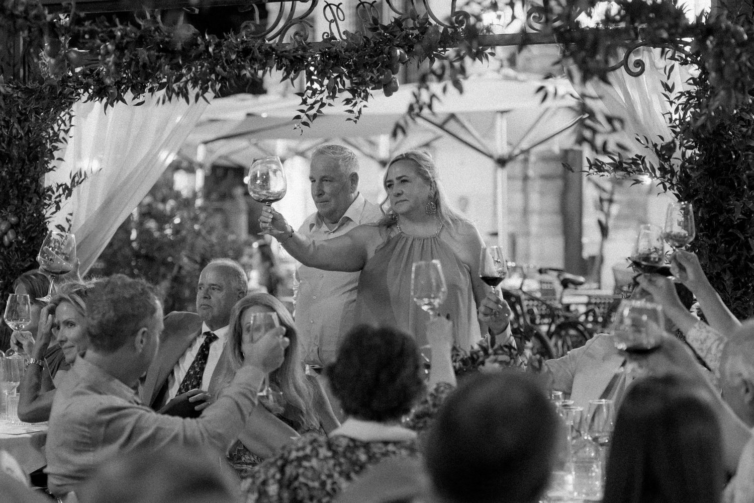 Black and white wedding reception toast with guests seated outdoors under floral arch decorations.