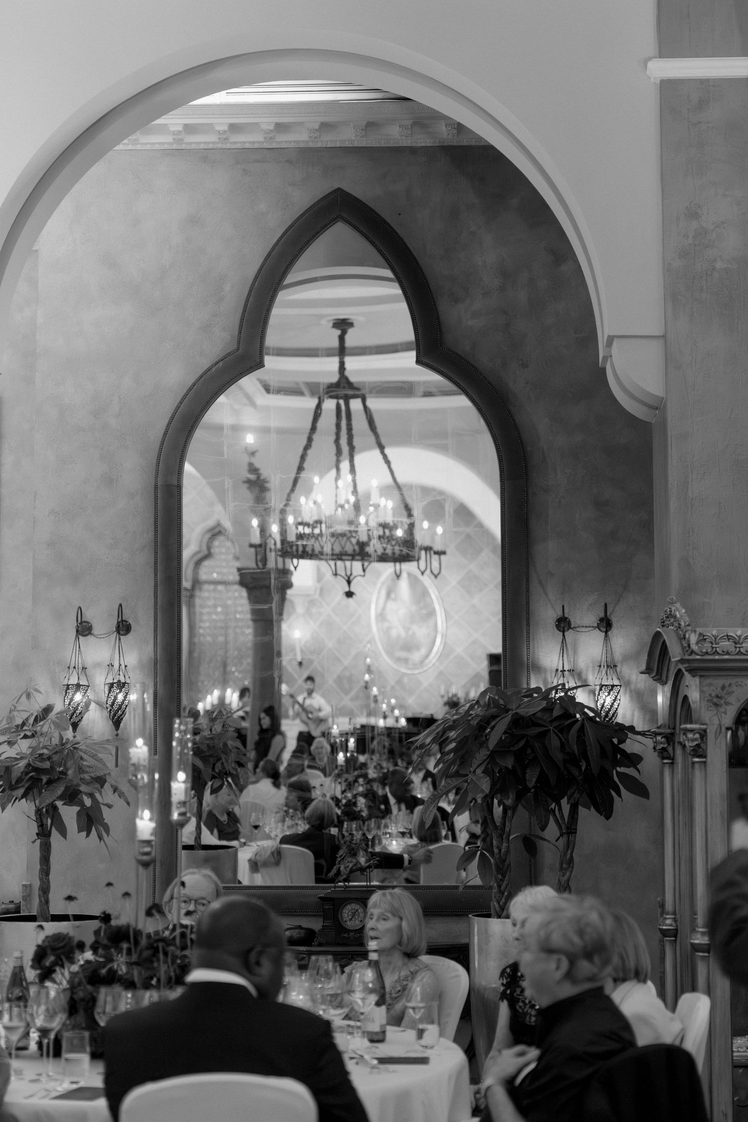 Black and white photo of wedding reception in elegant venue with Gothic arch, chandelier and guests dining at tables.