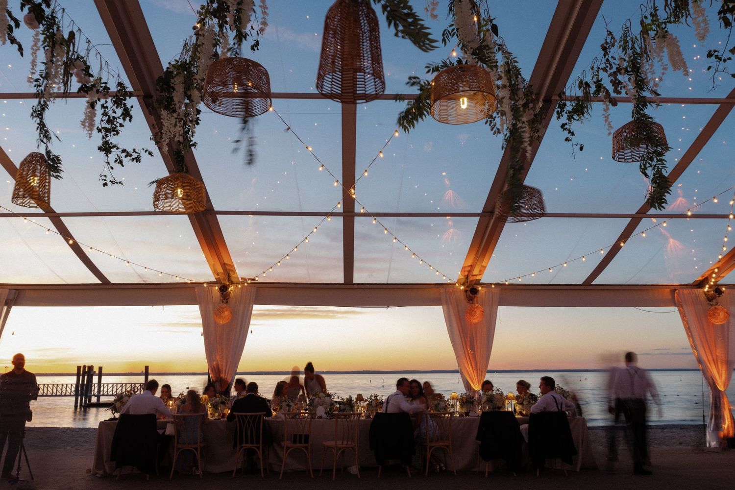 Stunning outdoor wedding reception under glass ceiling with string lights and ocean sunset views.