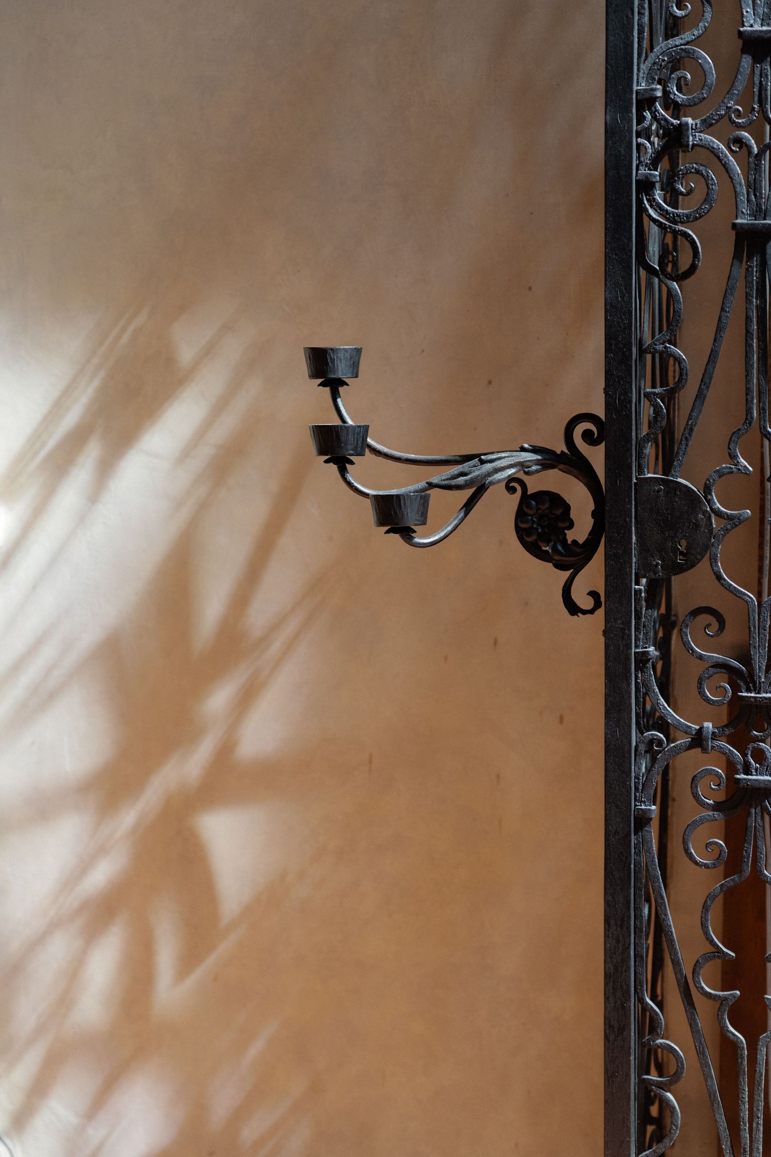 Ornate black wrought iron candelabra wall sconce beside decorative gate on warm beige stucco wall.