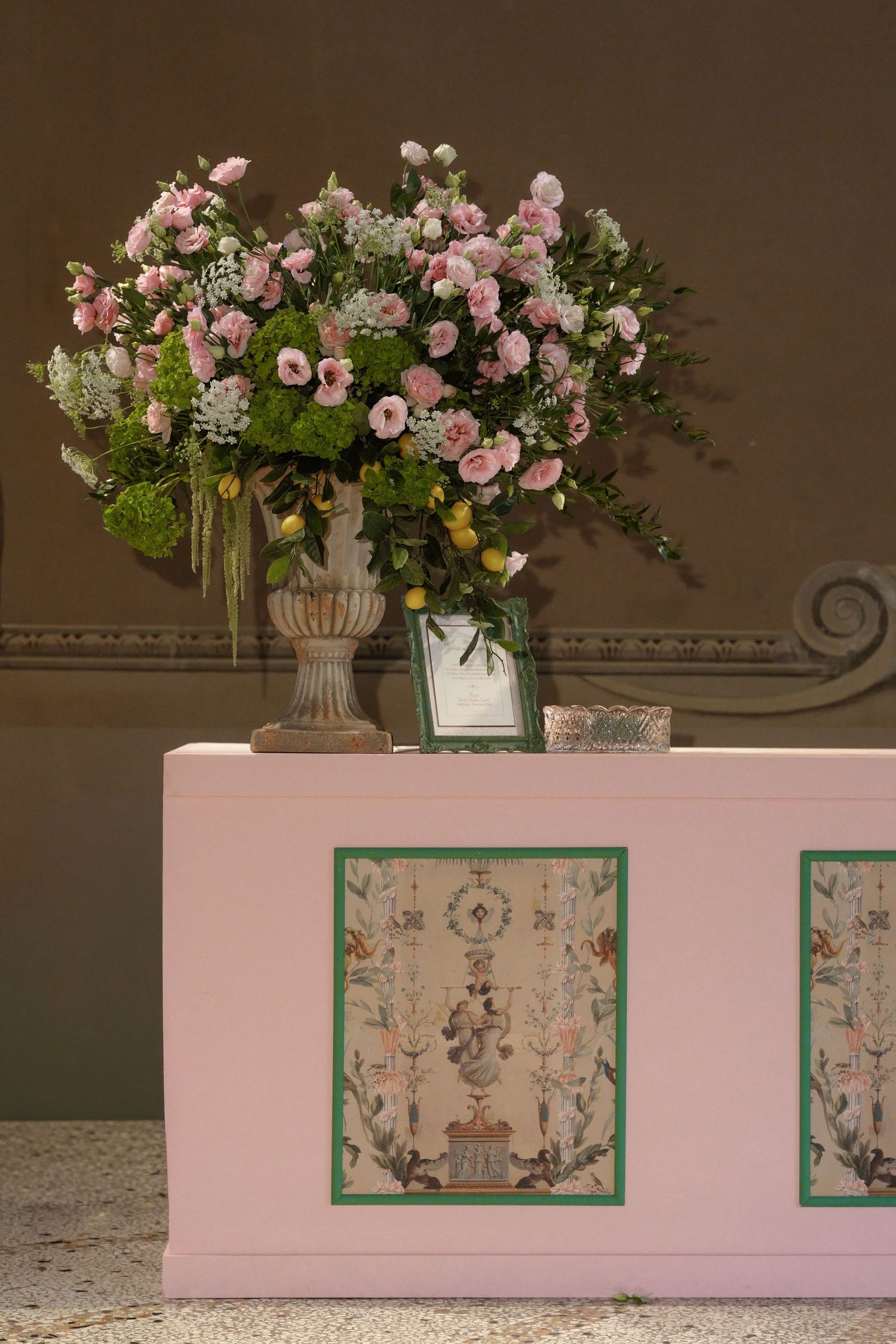 Pink floral arrangement in stone urn atop decorated pink cabinet with painted panels in elegant interior setting.
