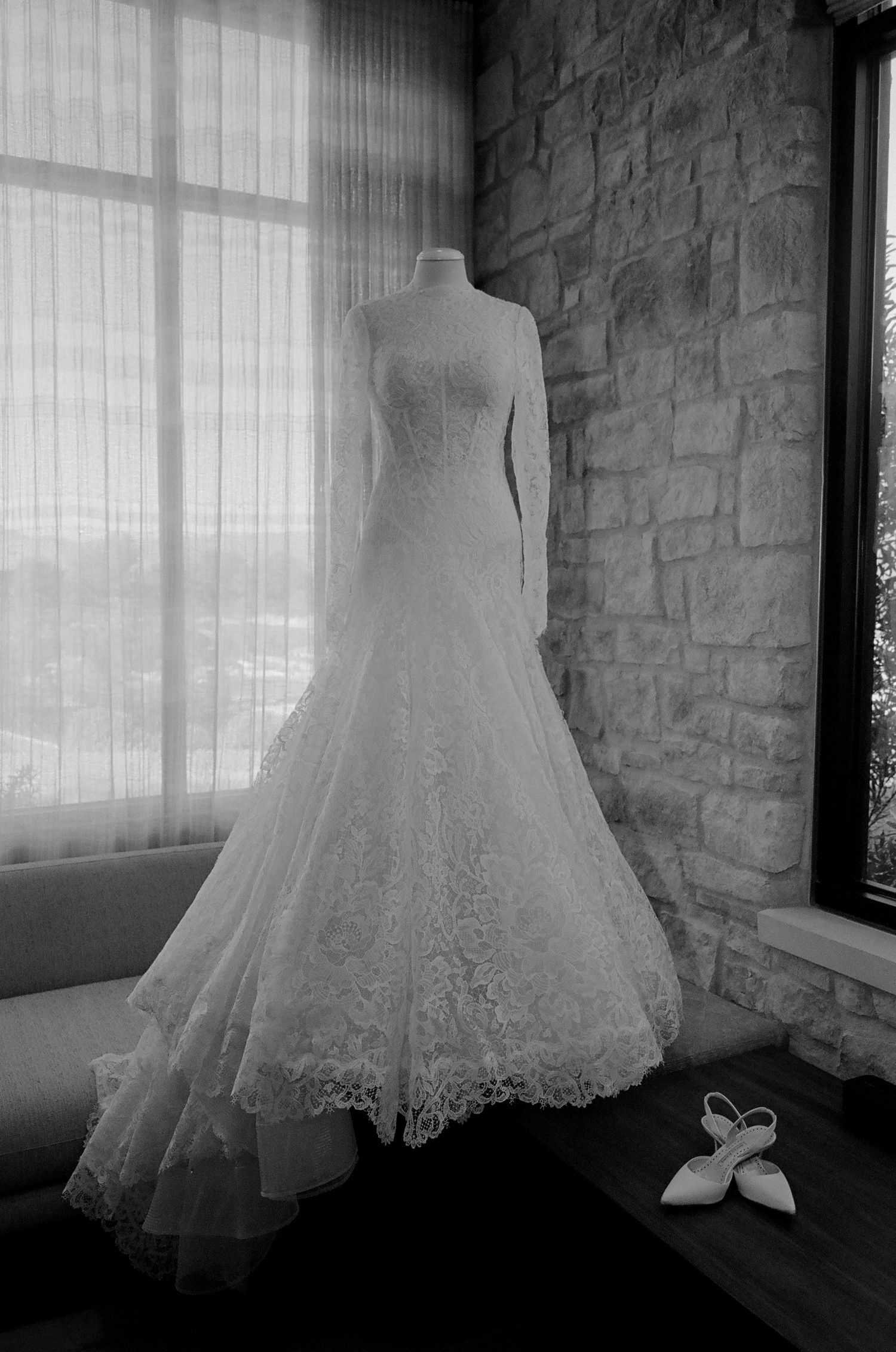 Elegant lace wedding dress displayed on mannequin against stone wall with white shoes.