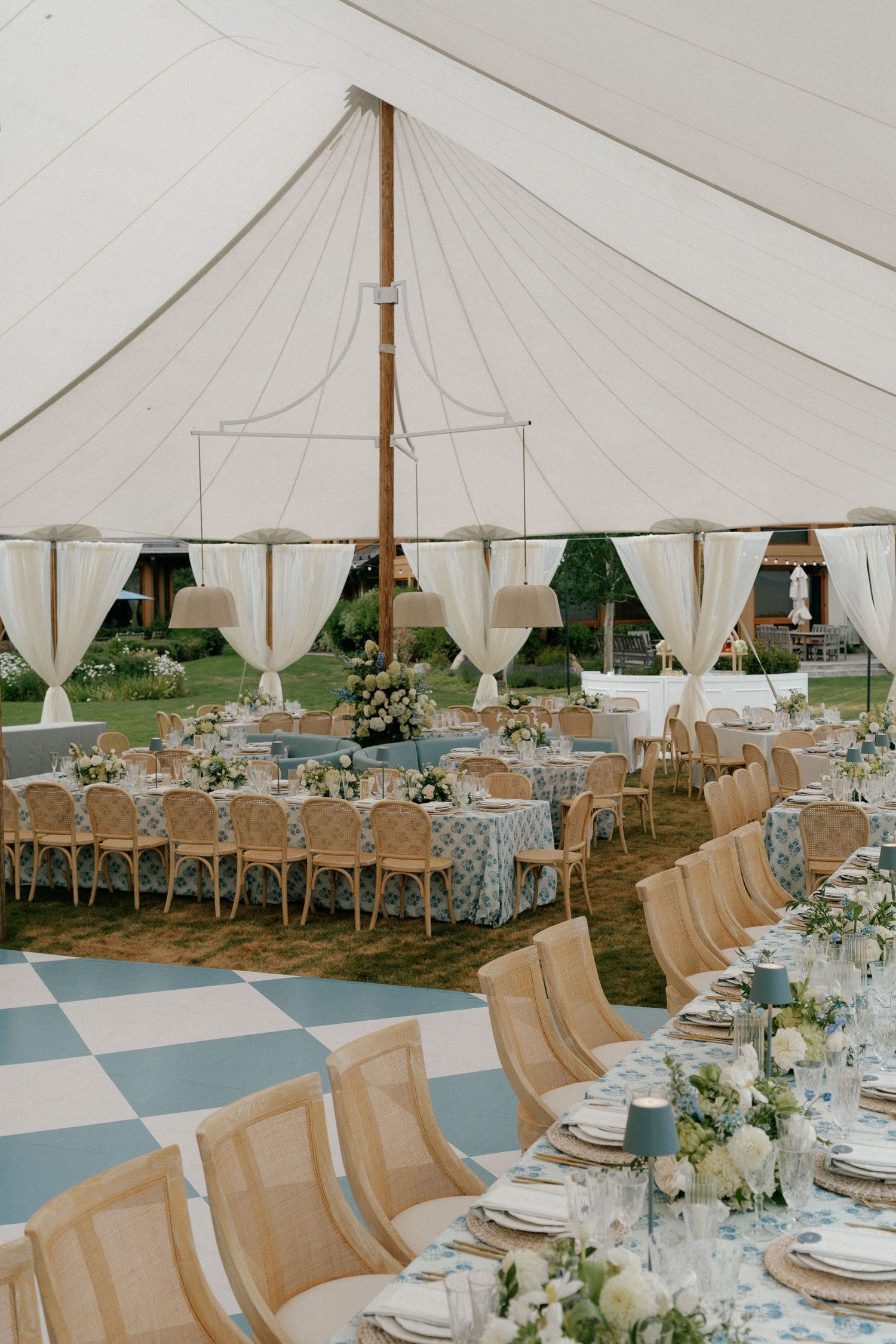 Interior of a white wedding tent with elegant table settings, cross-back chairs, and floral centerpieces.