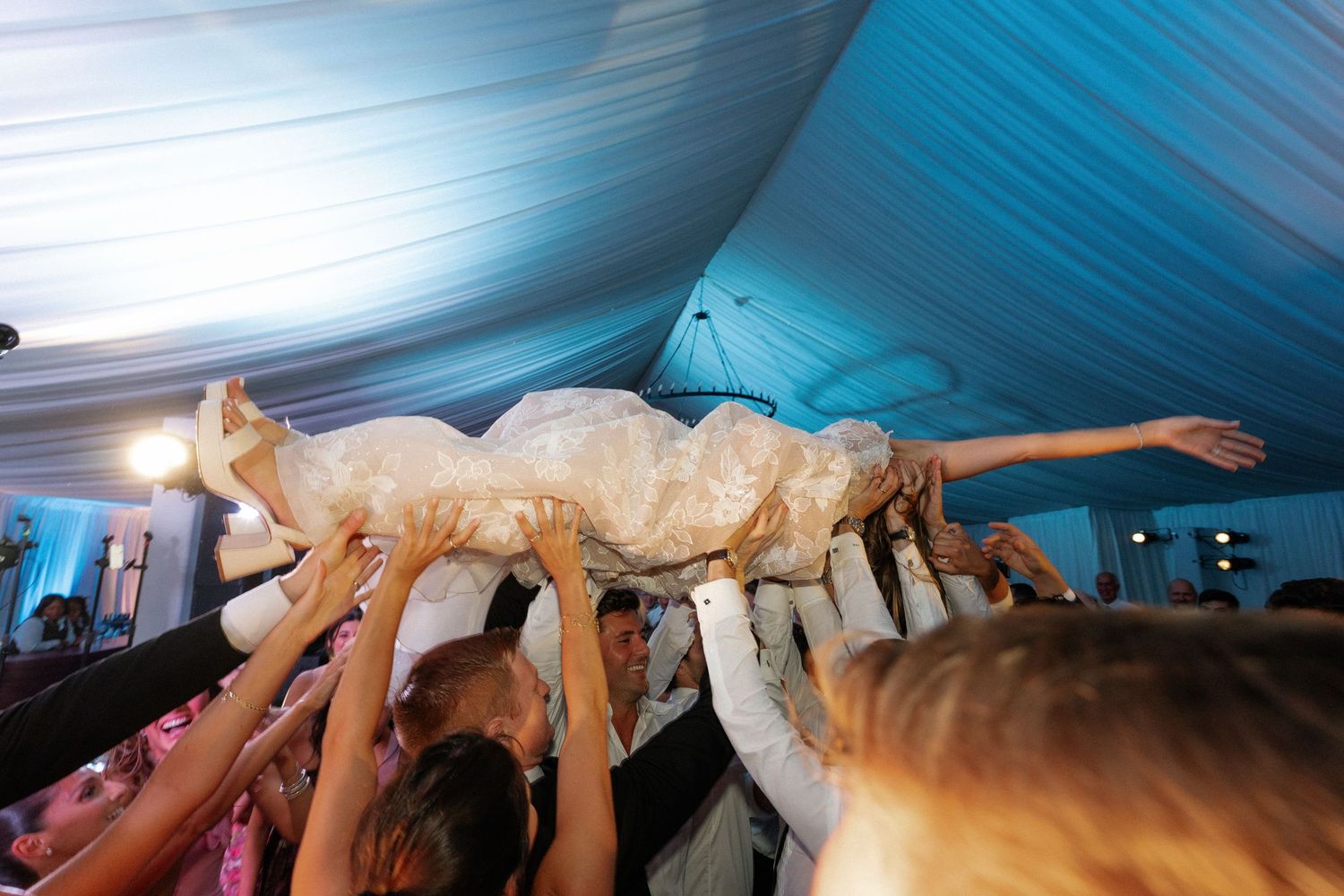 Dramatic dance move with bride being lifted horizontally under a blue-lit tent ceiling.