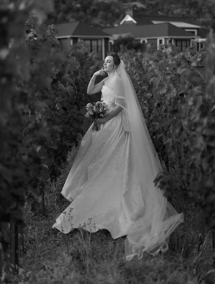 Bride in flowing white gown posing among garden flowers in black and white photography.