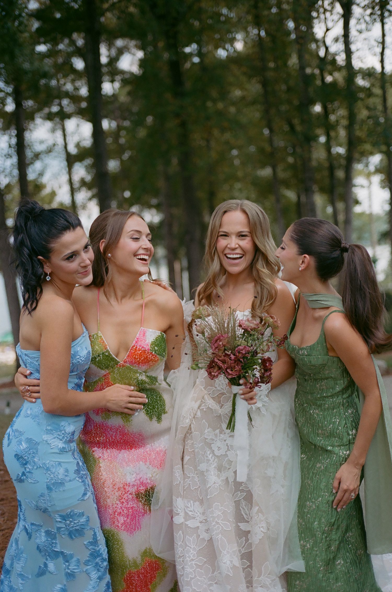 A group in colorful formal dresses share joyful laughter while holding bouquets in a wooded outdoor setting.