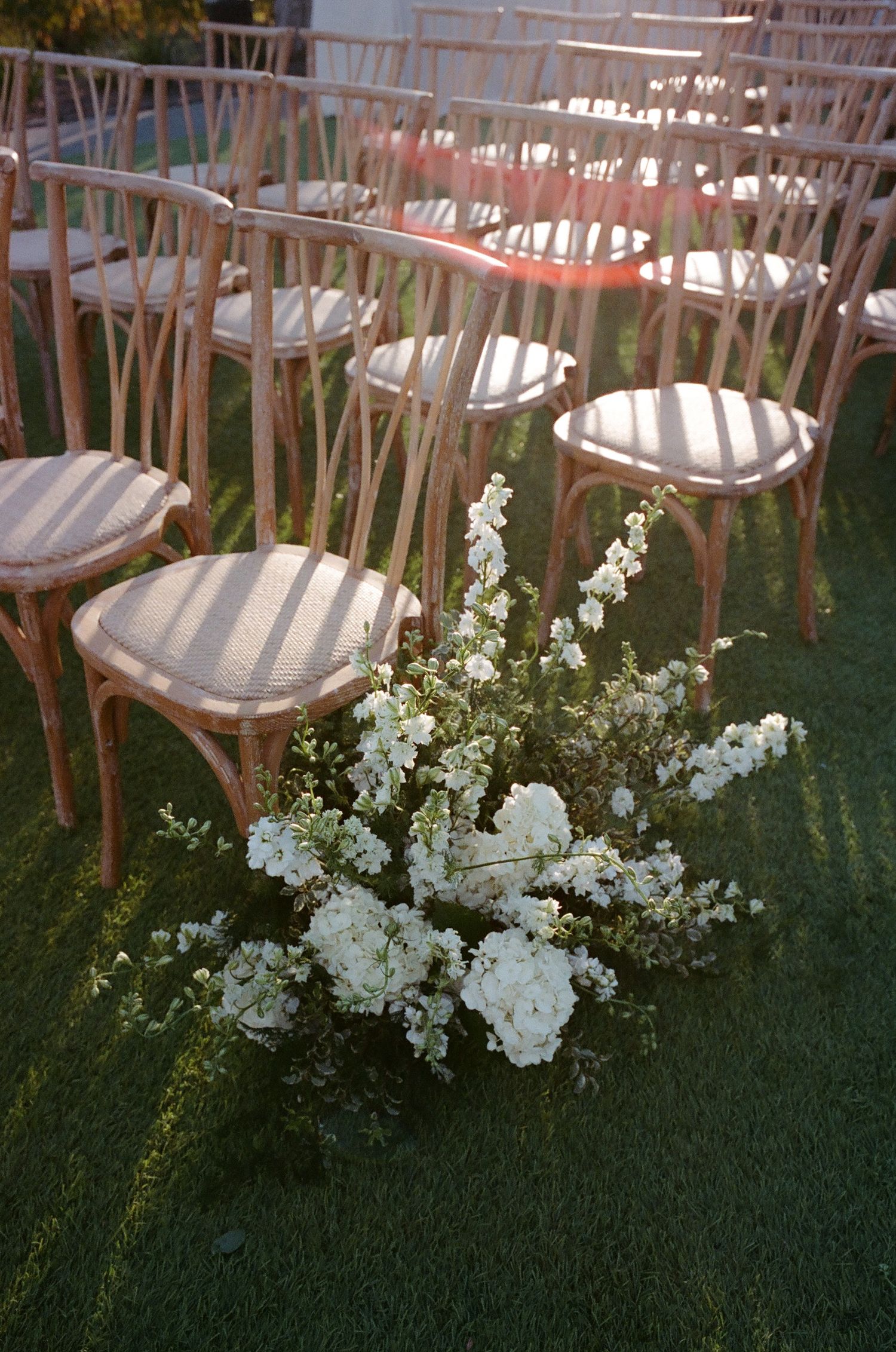 White wooden ceremony chairs lined up with delicate white floral arrangements along the aisle.