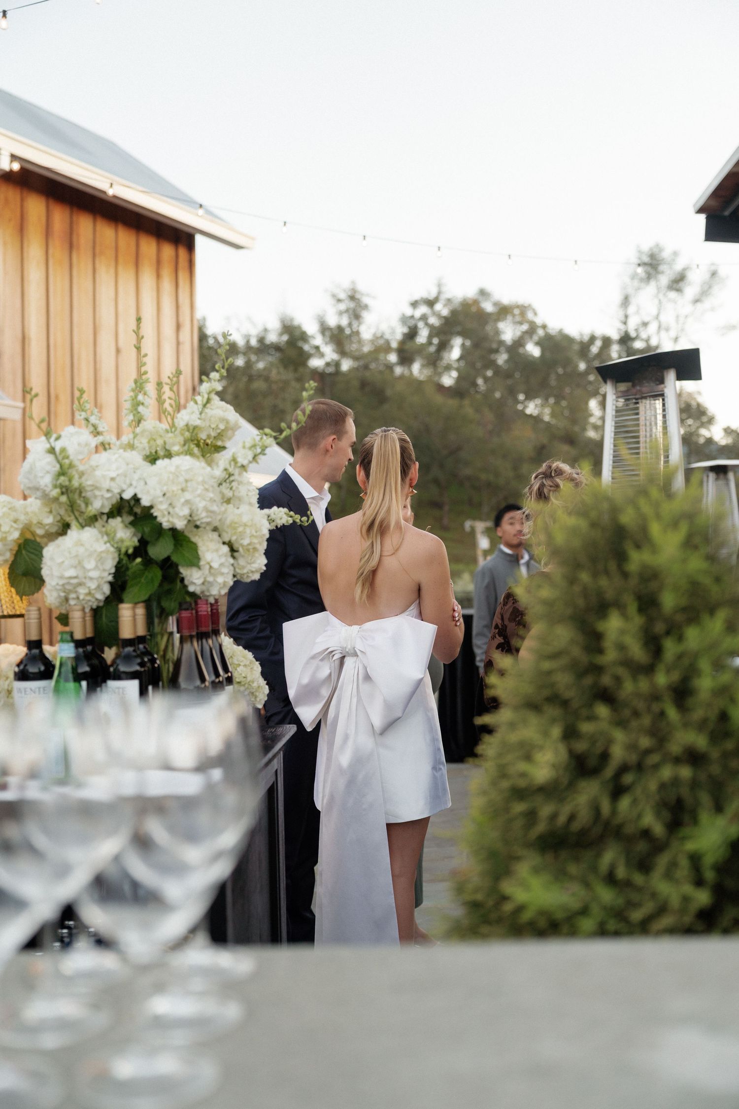 Elegant outdoor wedding reception setup with white floral arrangements and greenery against a rustic wooden barn backdrop.