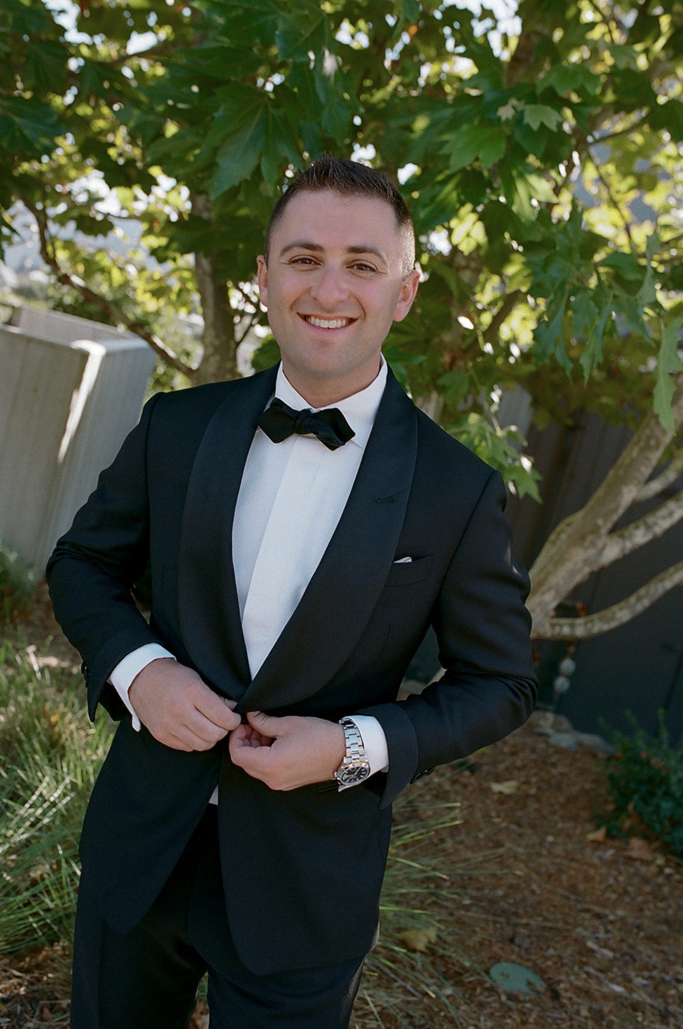 Wedding party member adjusting formal black tuxedo outdoors under tree canopy.