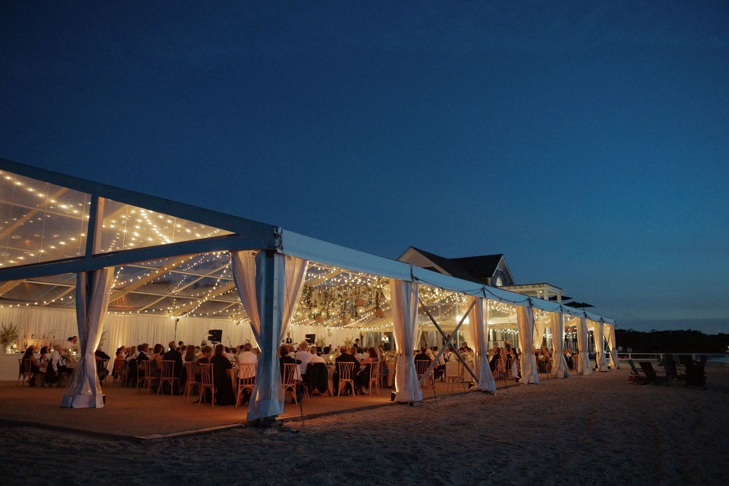 Elegant beachside wedding reception under white tent with string lights at dusk.