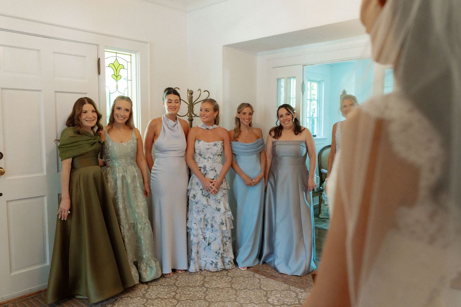 Bridesmaids in sage green and blue dresses react emotionally during a first look moment with the bride in a white gown.