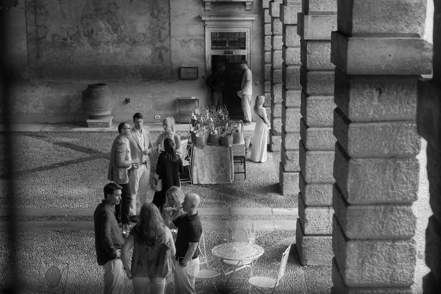 Black and white photo of elegantly dressed guests socializing under grand stone archways at a formal outdoor event.