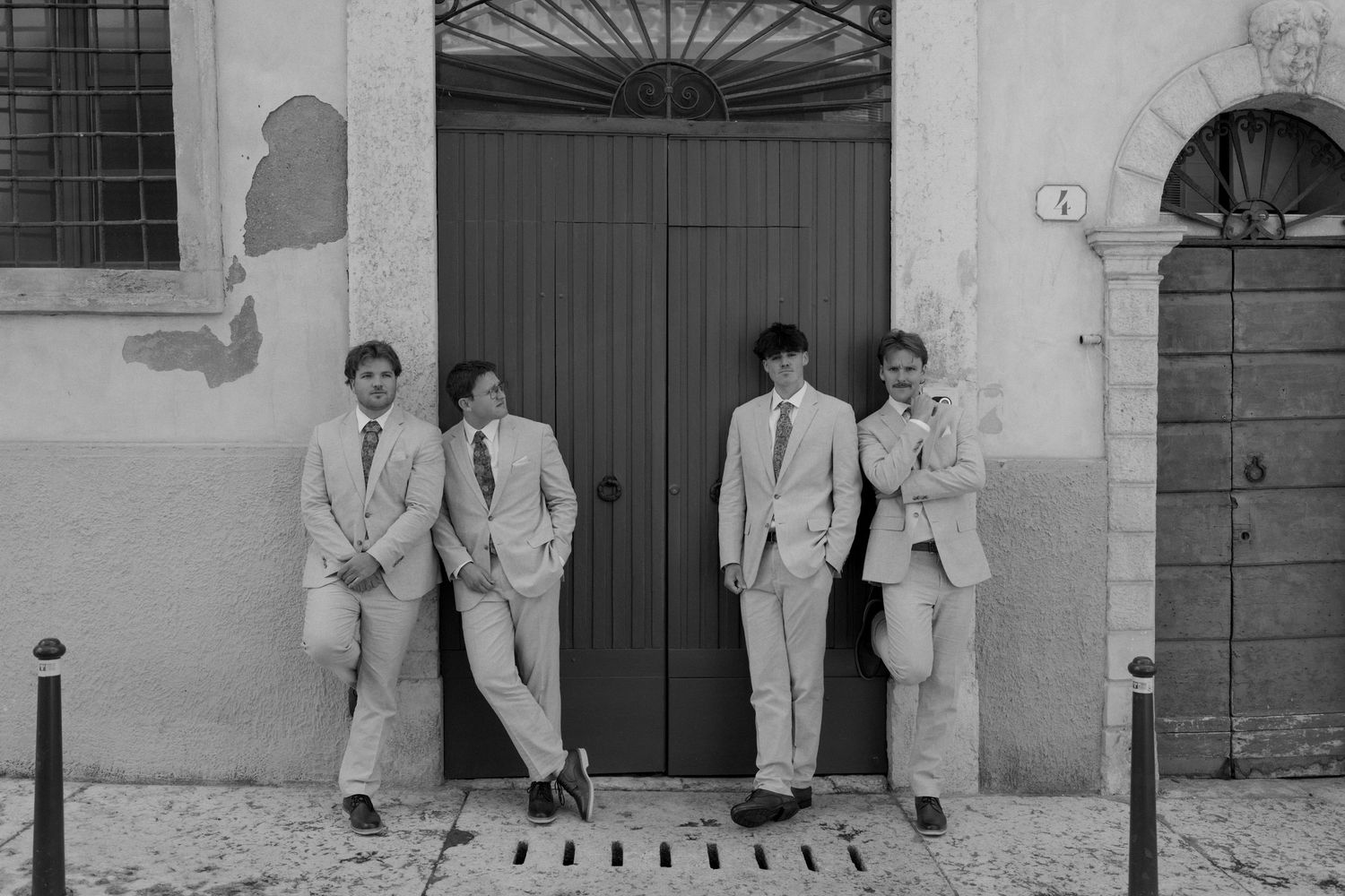 Four young men in suits lean against a weathered building facade with a green door in a black and white photo.