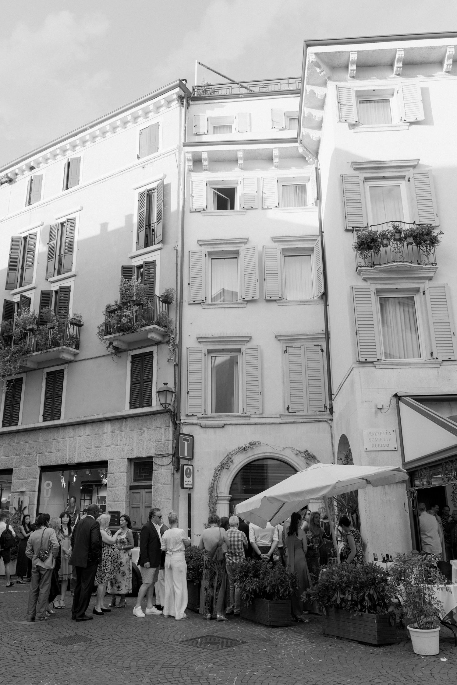 Black and white photo of wedding guests mingling outside a charming Italian building with shuttered windows and balconies.