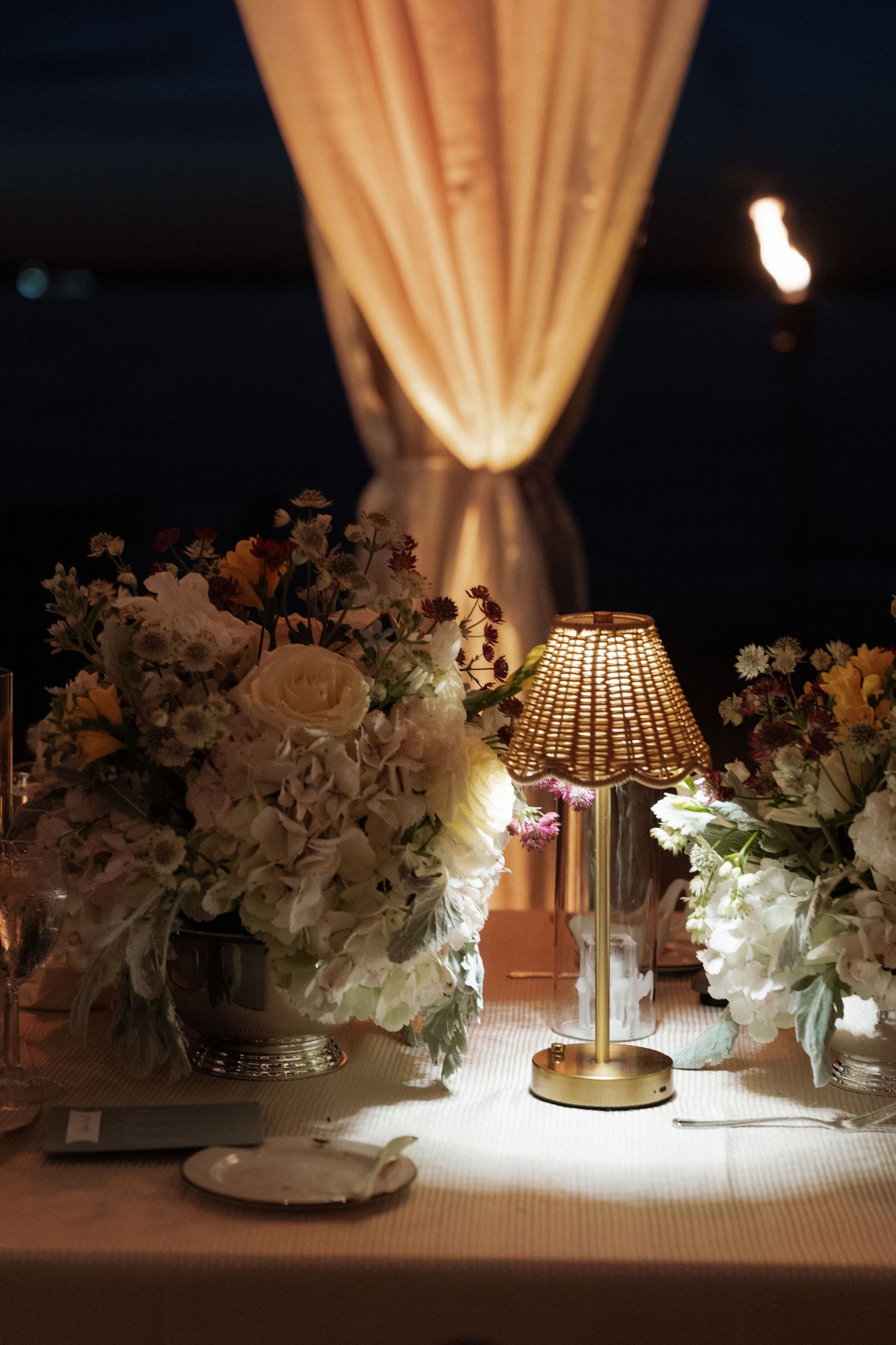 Elegant wedding table with glowing lamp, white floral arrangements, and draped curtains in romantic evening setting.
