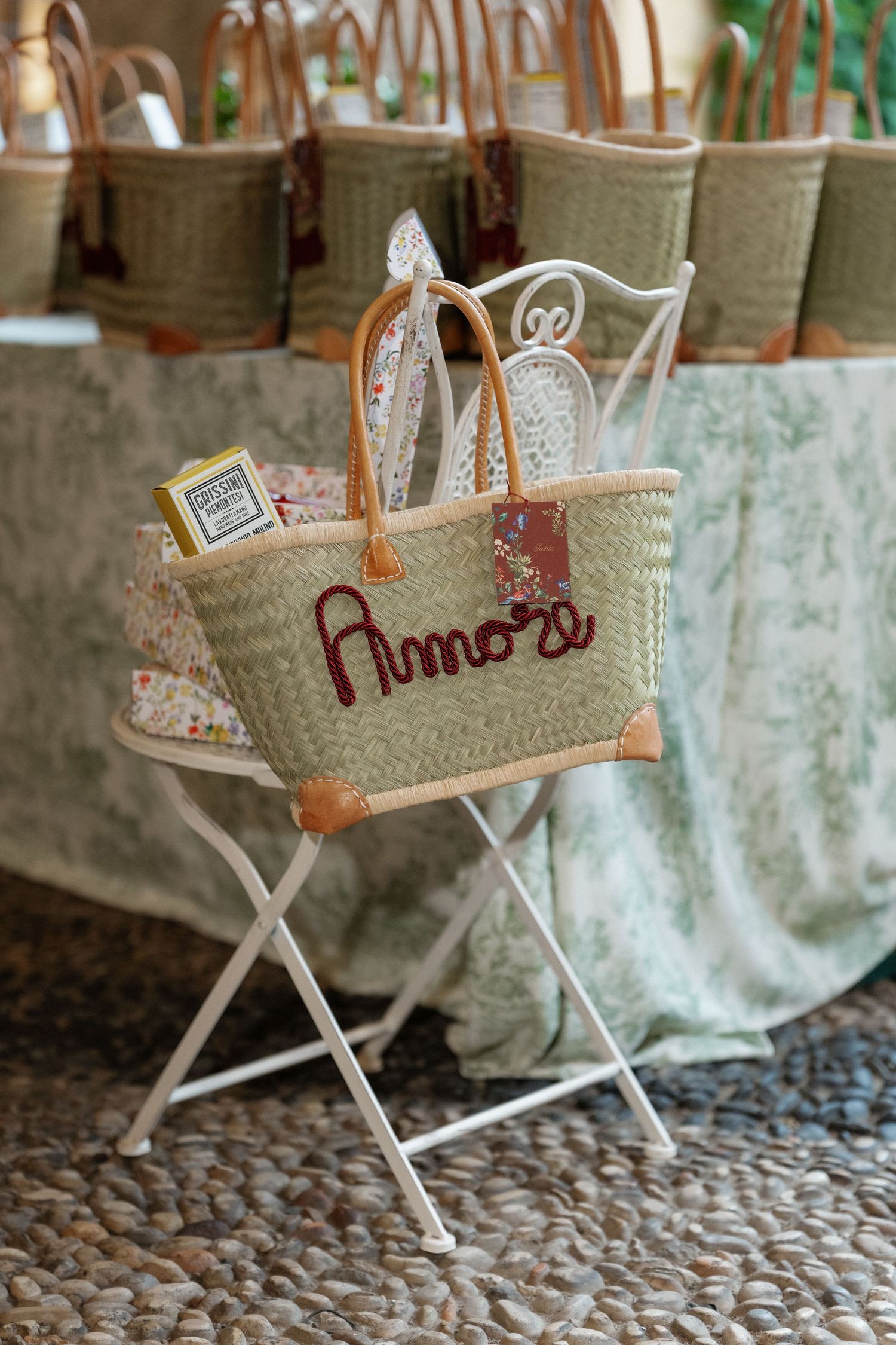Wicker tote bag with leather handles, embroidered with Amore, displayed on a white metal chair at a wedding.