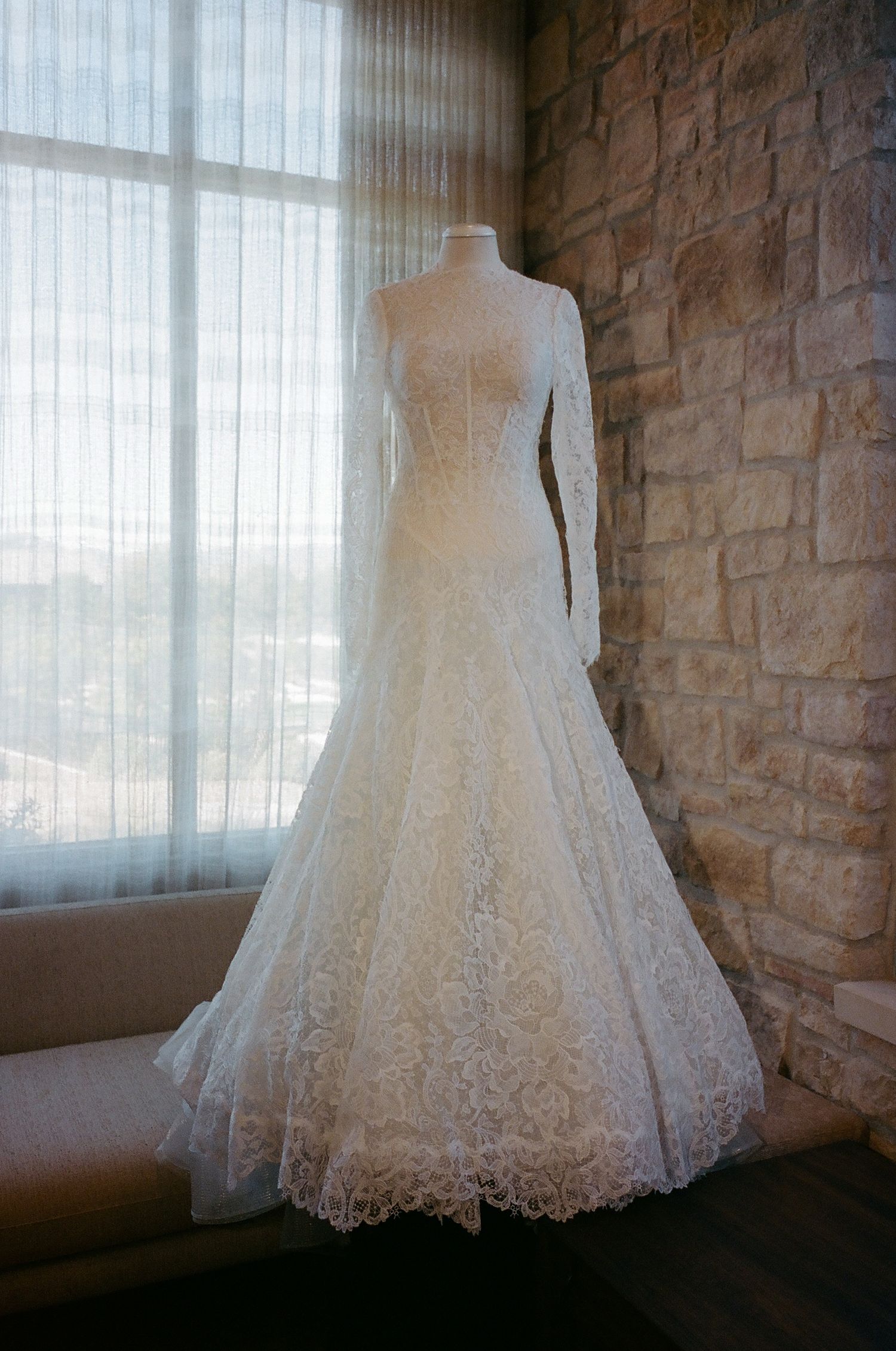 Elegant white lace wedding dress displayed against stone wall with natural window light.