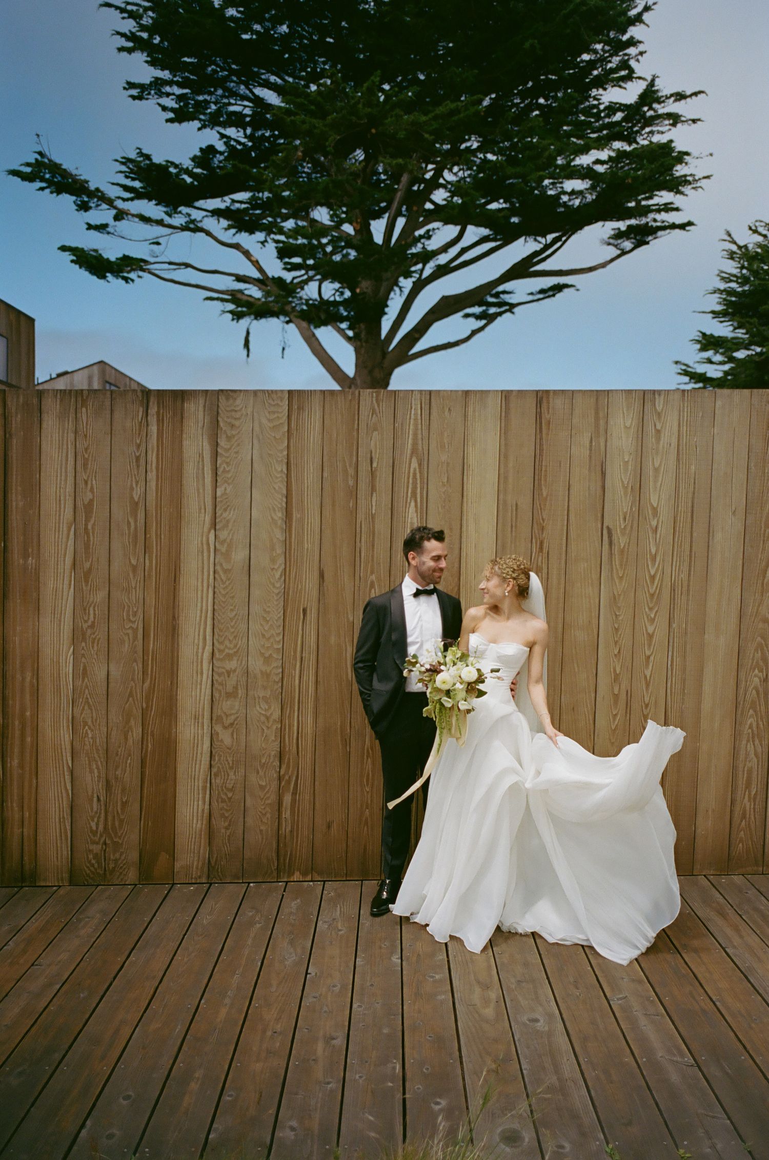 A couple poses playfully on a wooden deck against a rustic fence for wedding photos.